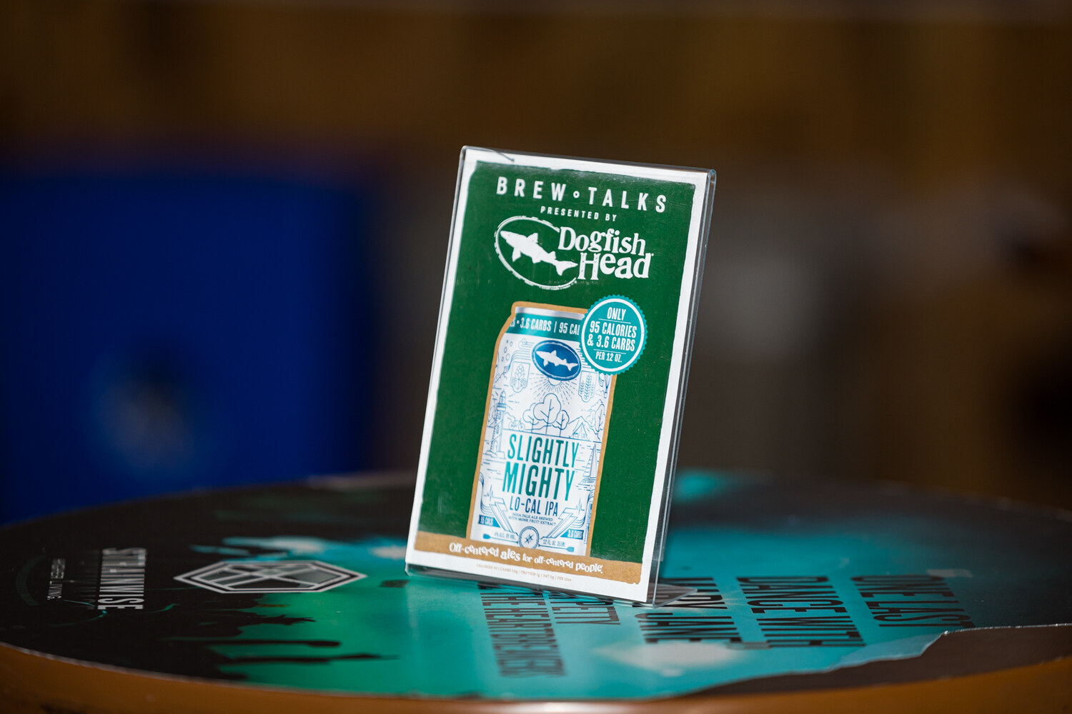 A green Brew Talks table card showing a Dogfish Head Slightly Mighty can on a dark wooden table.