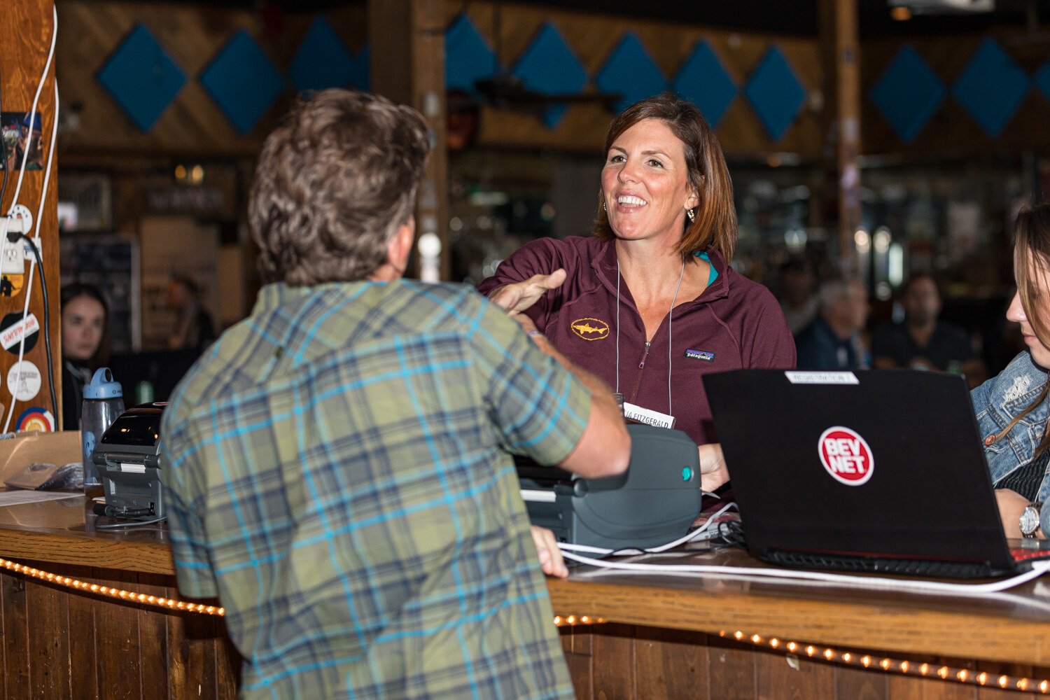 A woman in a maroon pullover behind a wooden counter greeting a man in a plaid shirt, with a label printer and laptop on the counter.