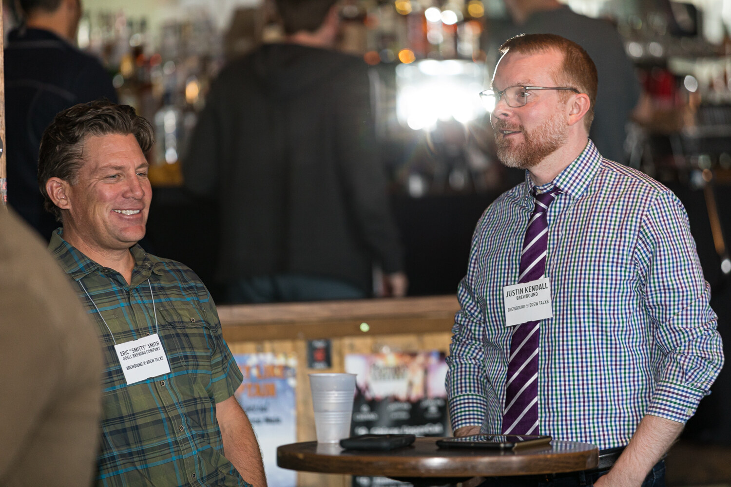 Two men with name badges laughing in conversation beside a high table, a crowded bar behind them.