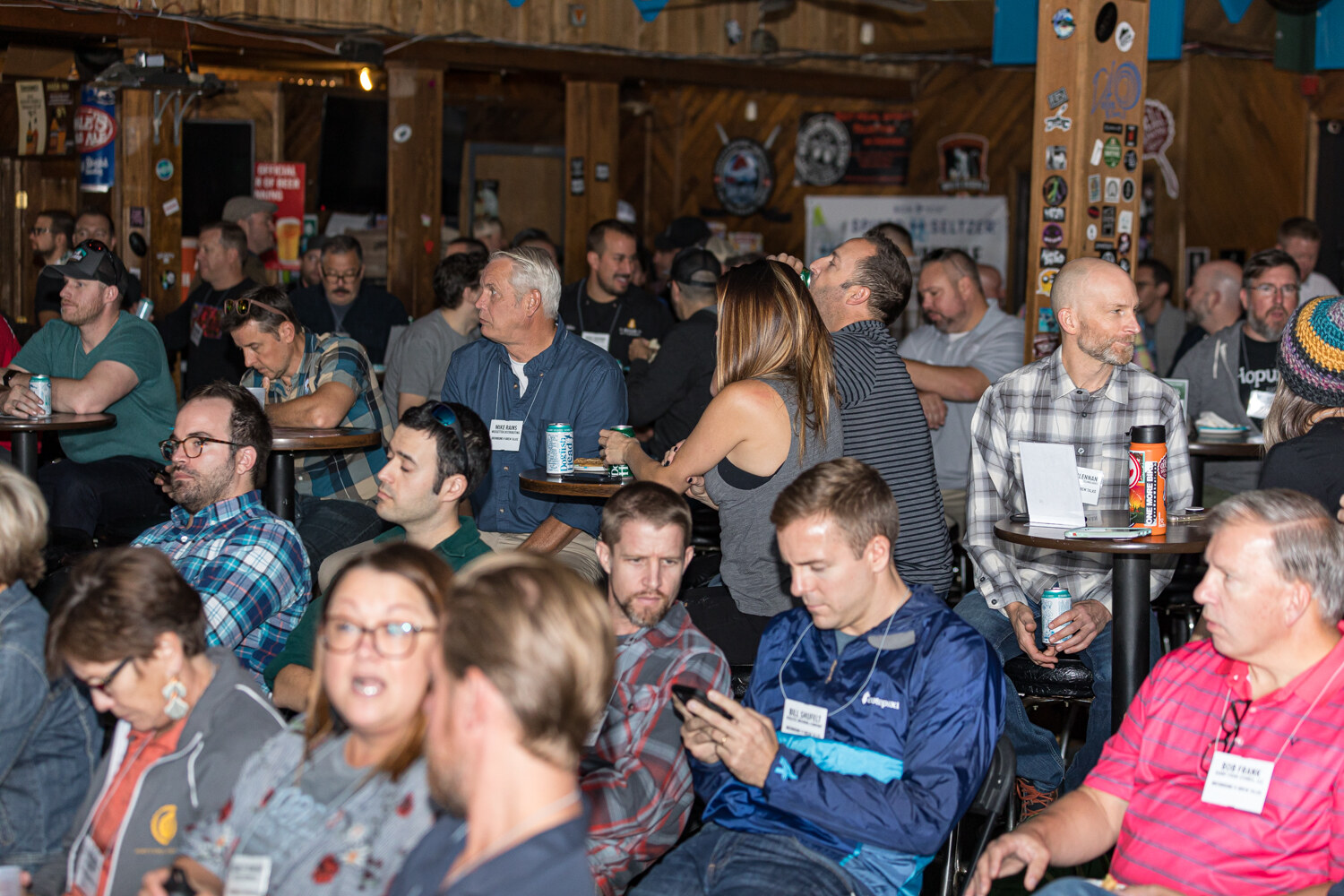 Crowd of seated attendees at small tables in a wood-paneled bar, many holding cans and wearing name badges.