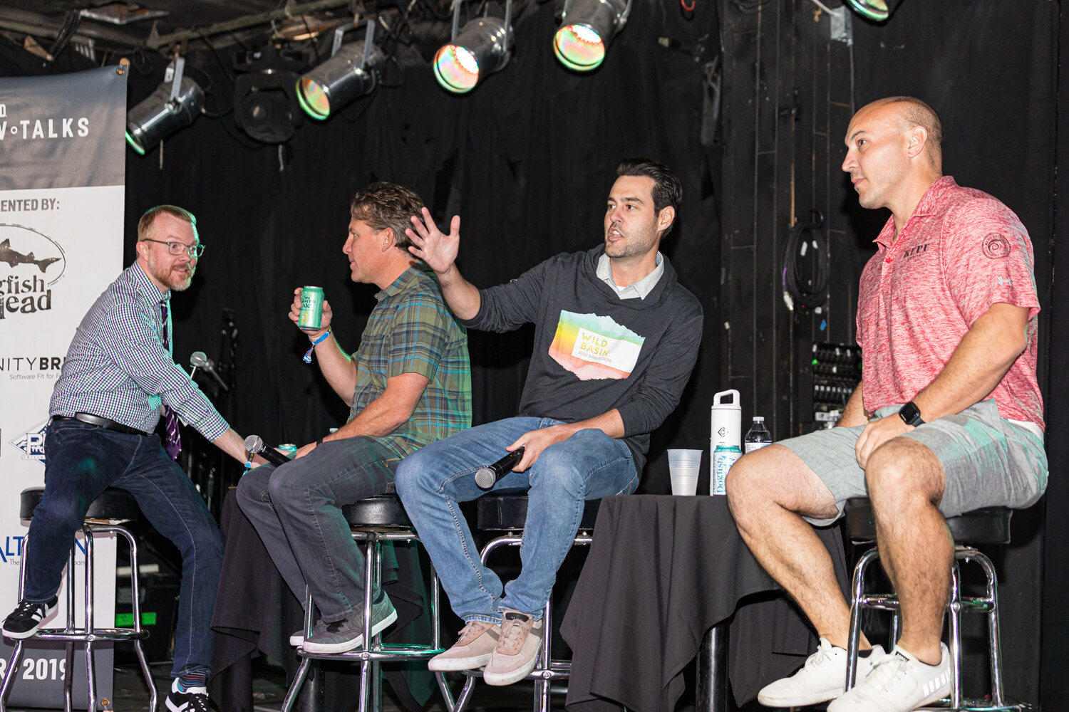 Four men seated on stools on a small stage with black curtains and Brew Talks banners.