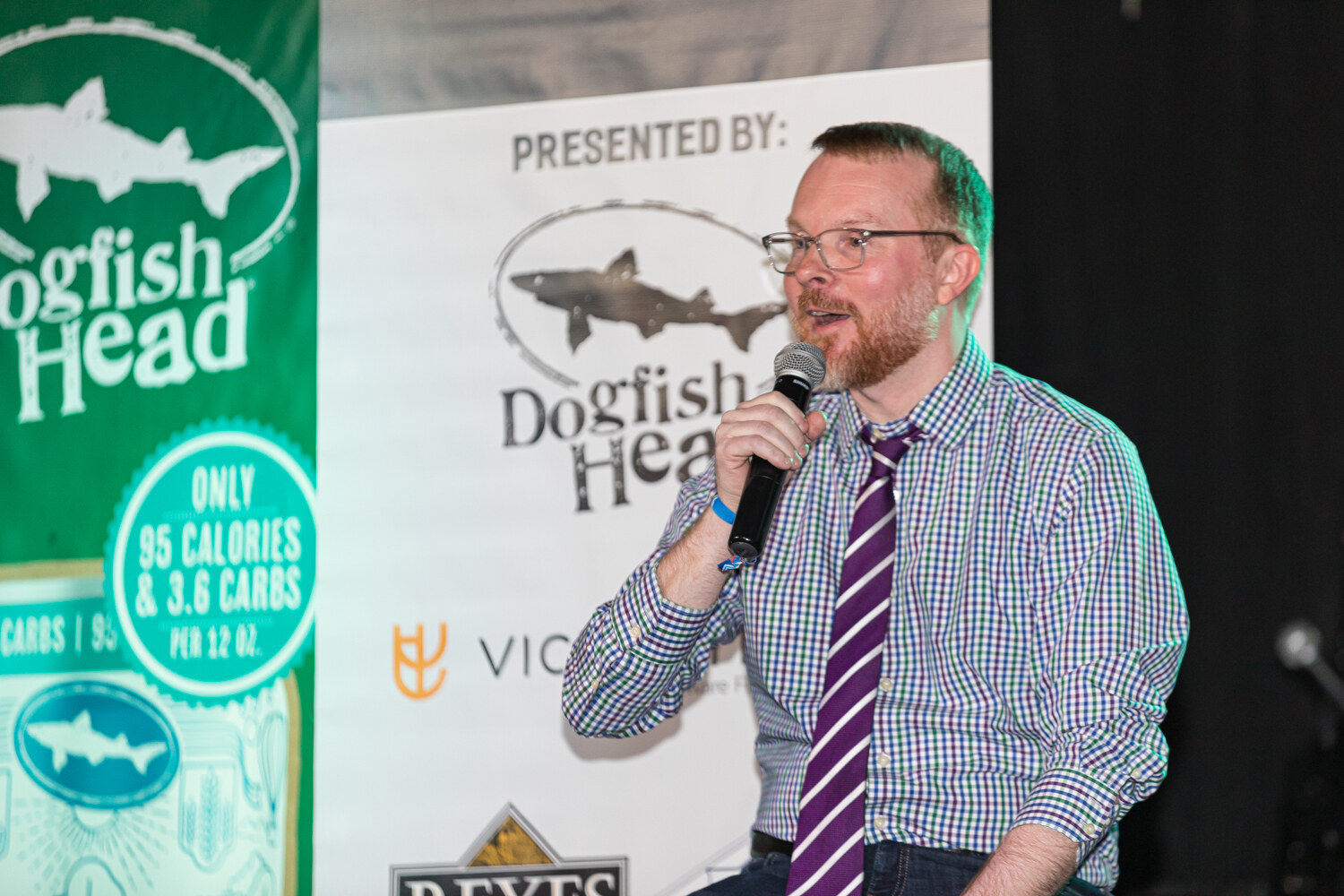 A man in a checked shirt and purple tie speaking into a microphone beside a banner reading Presented by Dogfish Head.
