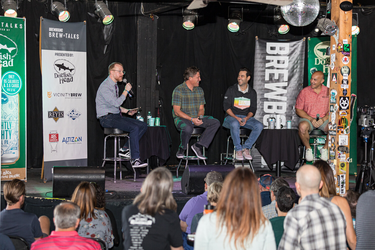 Wide shot of four people on stools on a small stage with a disco ball overhead and an audience in the foreground.