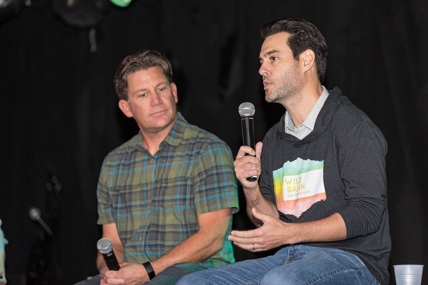 Two seated men, one in a green plaid shirt and one in a dark hoodie speaking into a microphone.