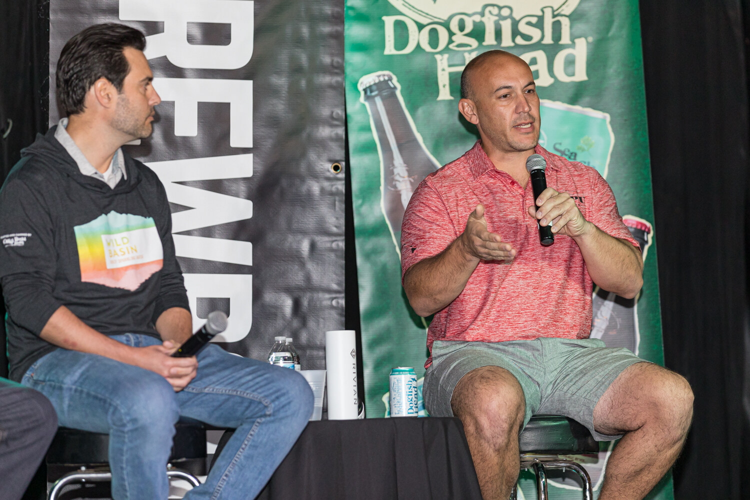 A man in a coral polo speaking with an open hand beside another panelist, with a green Dogfish Head banner behind.