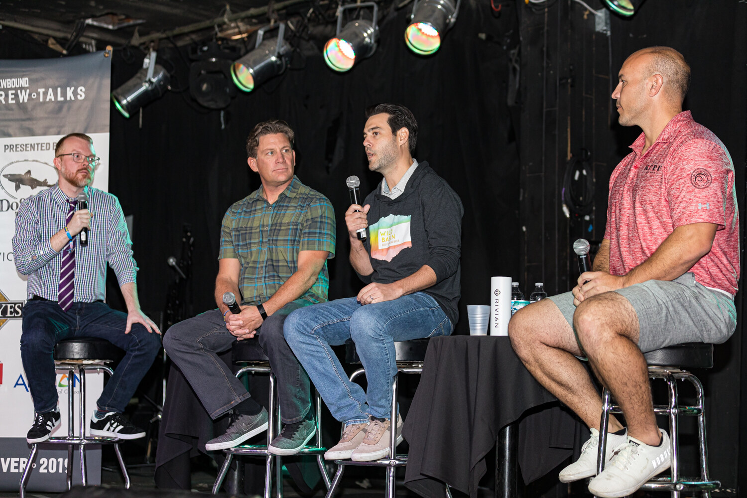 Four men on stools on a stage with a Brew Talks banner at the left and stage lights above.