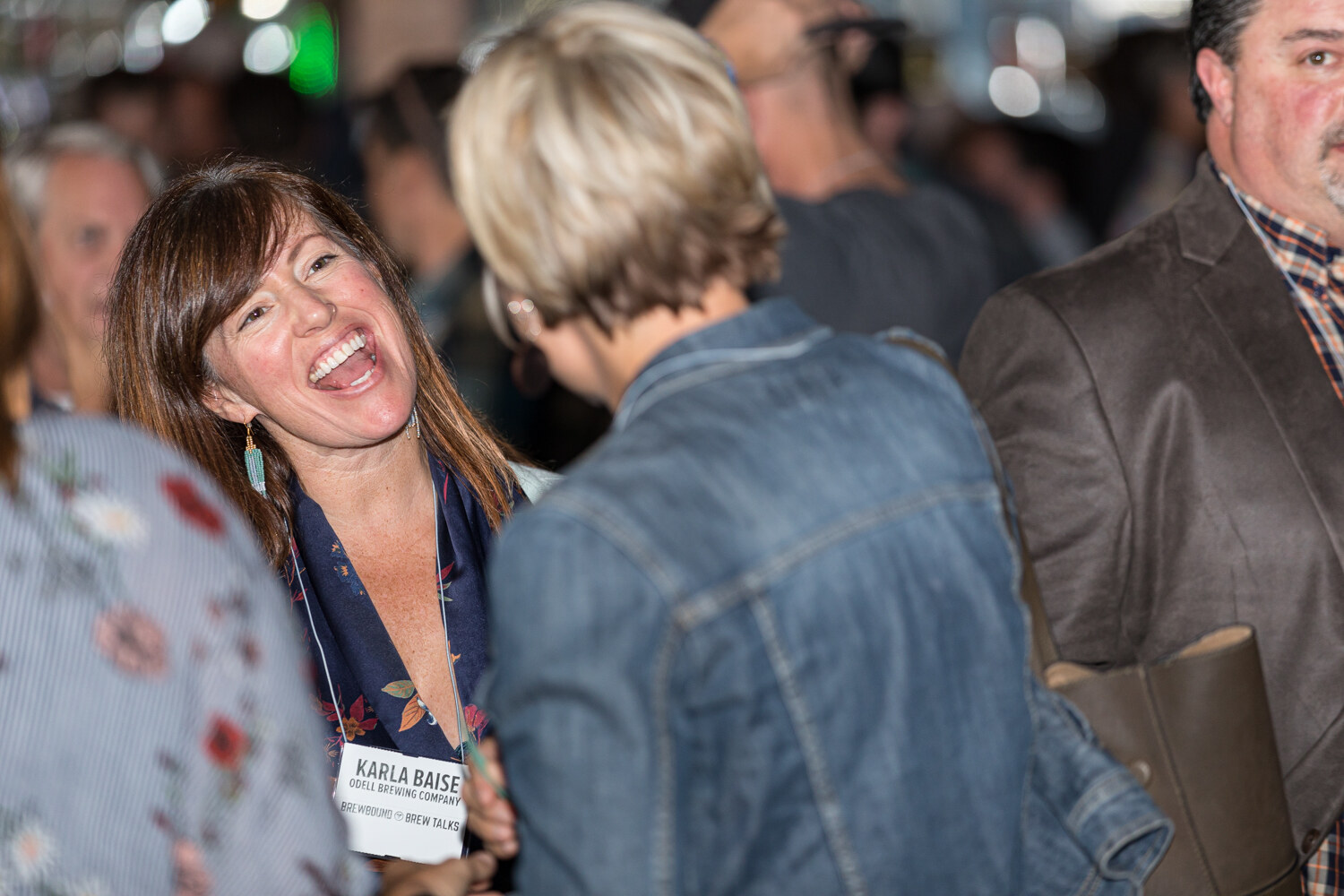 A woman with a lanyard laughing broadly in a busy room, other attendees around her.