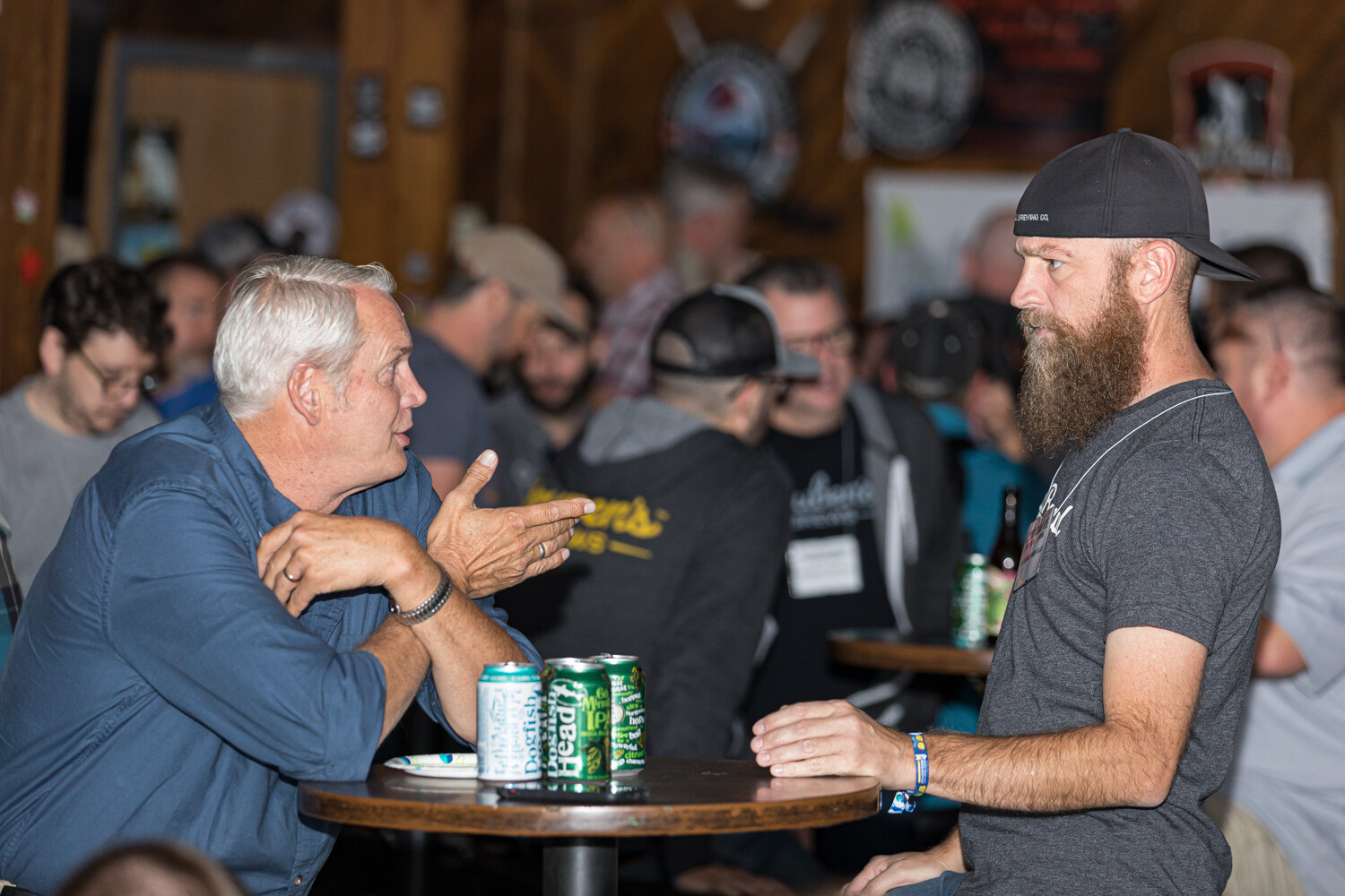 A white-haired man and a bearded man in a cap talking across a small round table with three beer cans.