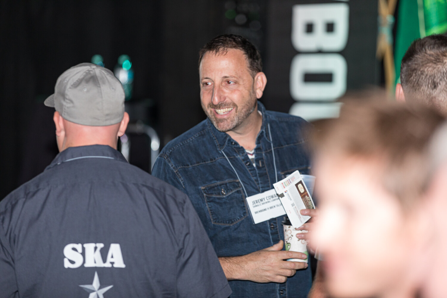 A smiling man wearing a name badge and a denim shirt, with another man in a gray cap in the foreground.