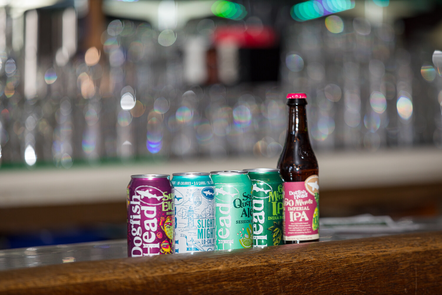Five Dogfish Head cans and a bottle lined up on a wooden bar in front of rows of glasses.