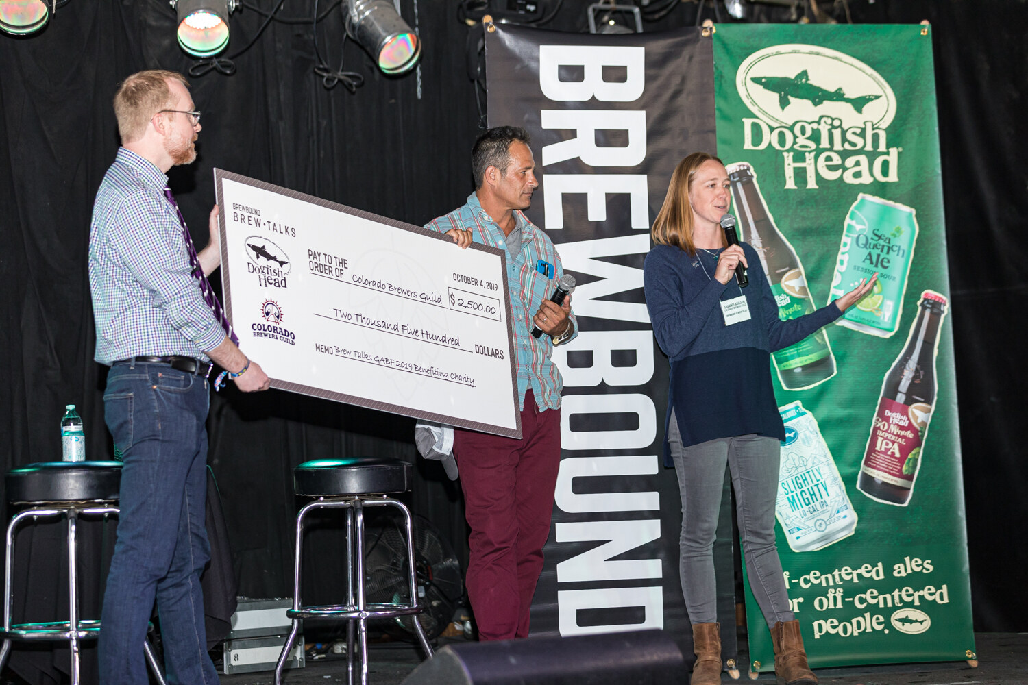 Three people on stage, two holding a large check dated October 4, 2019 made out to the Colorado Brewers Guild, in front of a Dogfish Head banner.