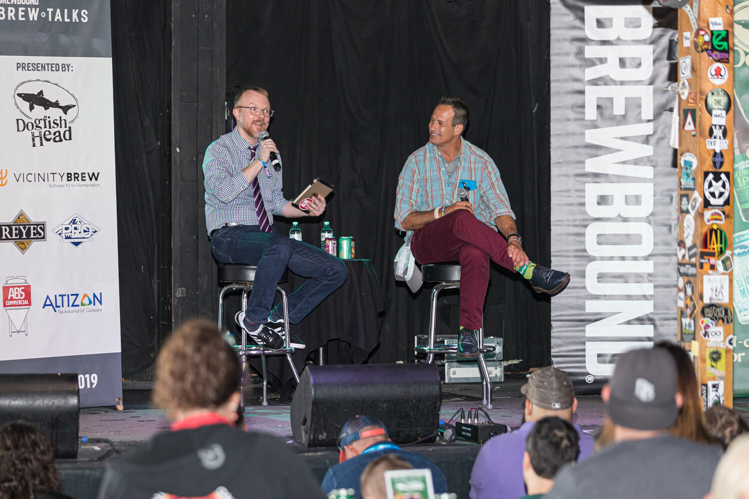 Two men seated on stools on a stage, one holding a tablet, with Brew Talks sponsor banners and an audience in front.