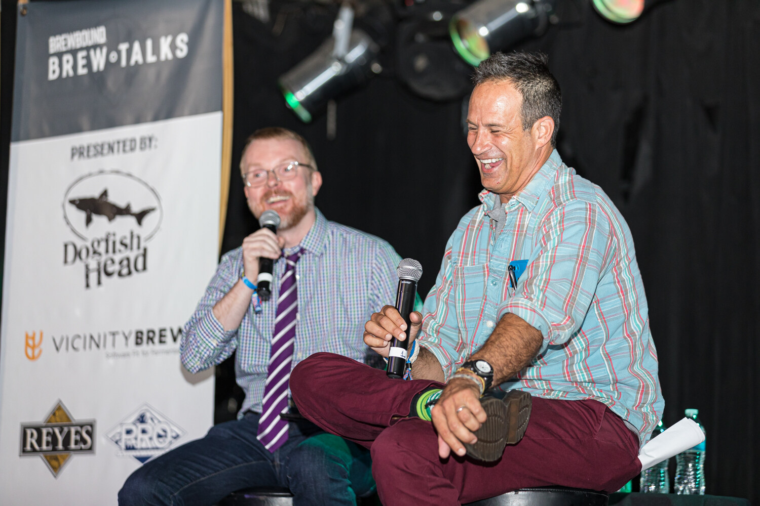 Two men laughing on stools, both holding microphones, beside a Presented by Dogfish Head banner.