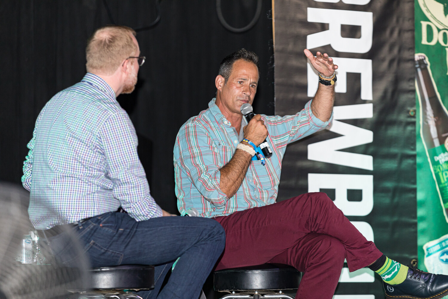 A man in a plaid shirt with a raised arm and microphone, the moderator seen from behind, with a Brewbound banner.