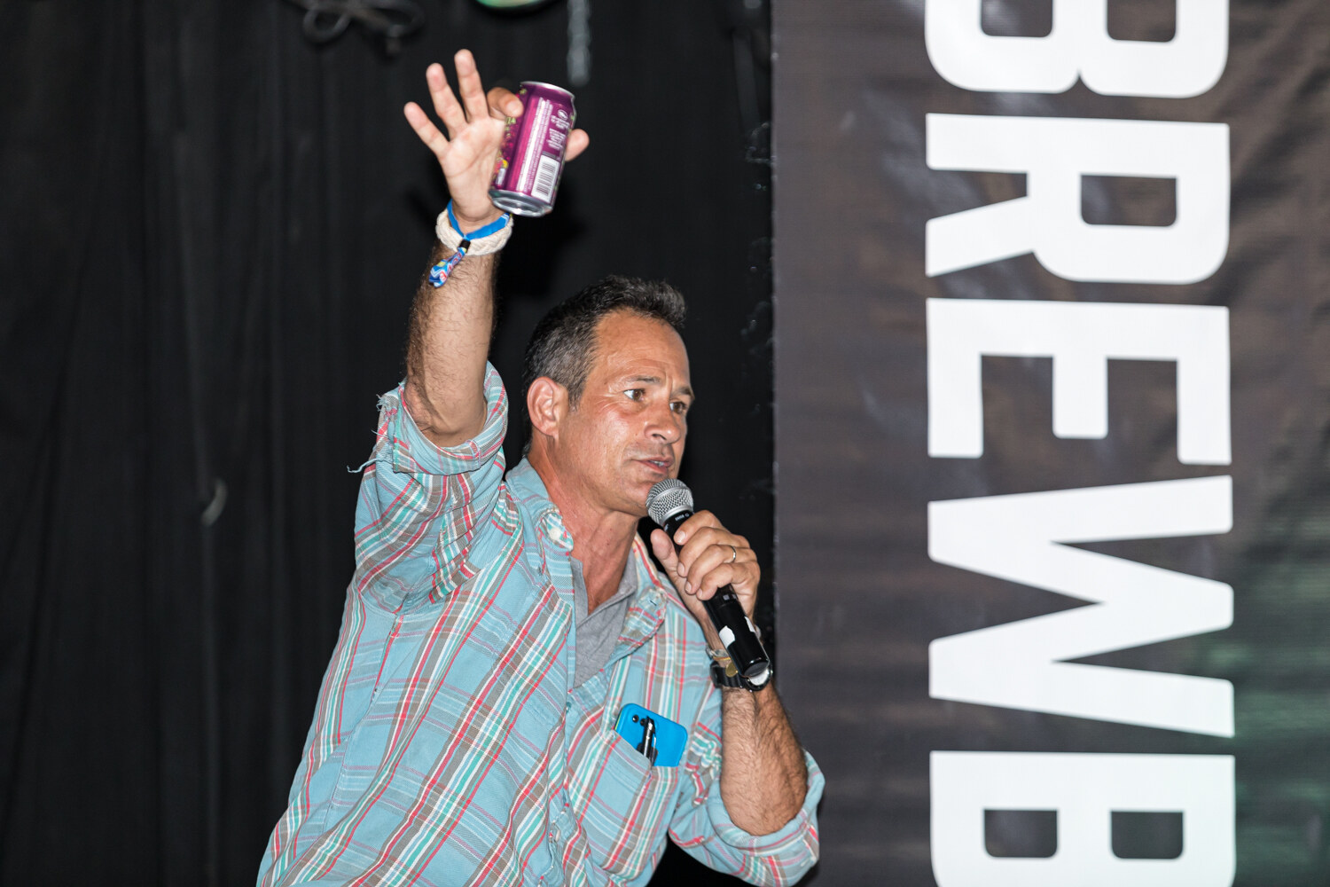 A man in a plaid shirt raising a purple can in one hand and holding a microphone in the other, against a Brewbound banner.
