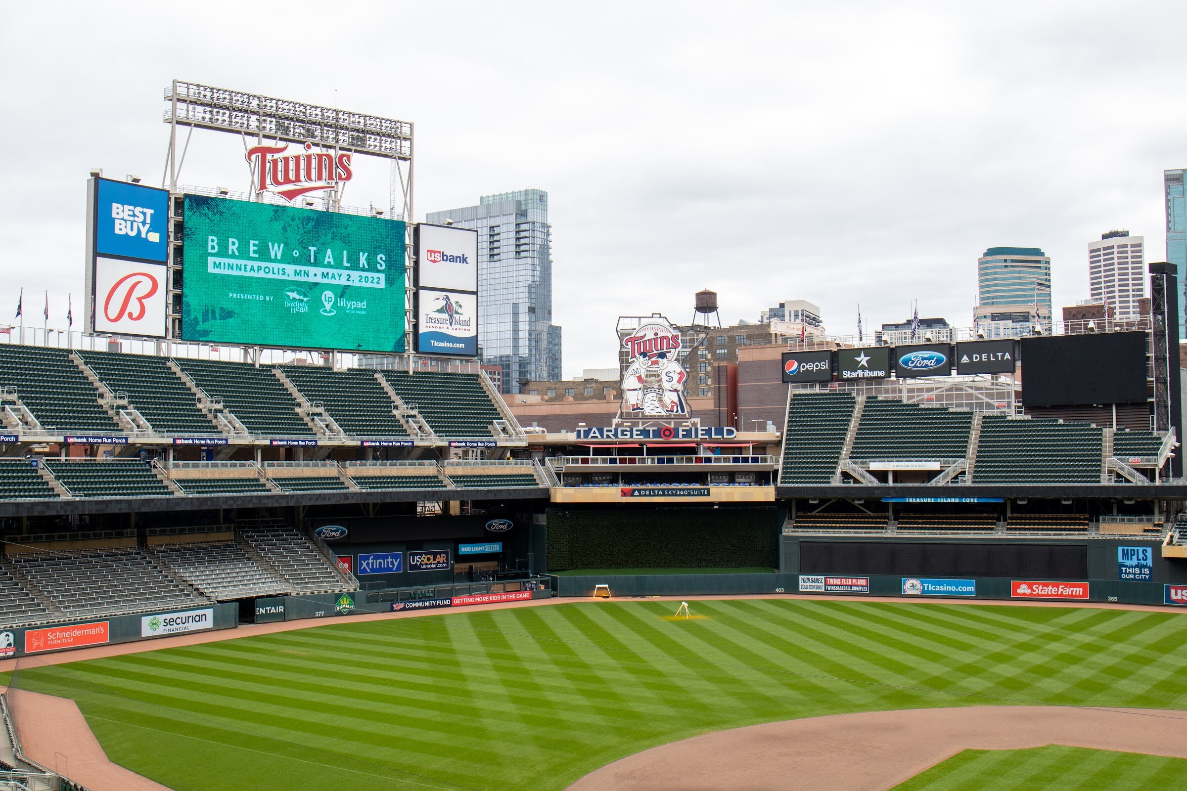 View across the Target Field infield to the Twins video board, where a green screen reads Brew Talks Minneapolis, MN, May 2, 2022 with sponsor logos, with the downtown skyline behind.