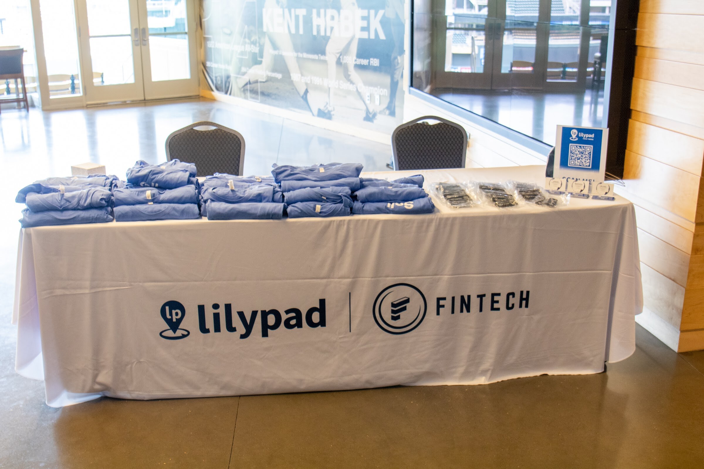 A white-skirted table labeled Lilypad Fintech holds stacks of folded blue shirts and bagged items, with a small Lilypad QR code sign at the right, in a concourse with a large Kent Hrbek photo wall behind.