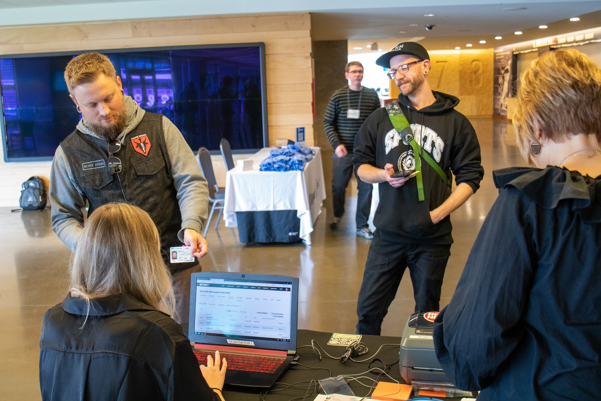 A bearded man in a gray hoodie and black vest holds up an ID card to a seated woman at a laptop with a badge printer, while a man in a black hooded sweatshirt and cap with a green strap stands behind, and a woman in a navy jacket is at the right edge.