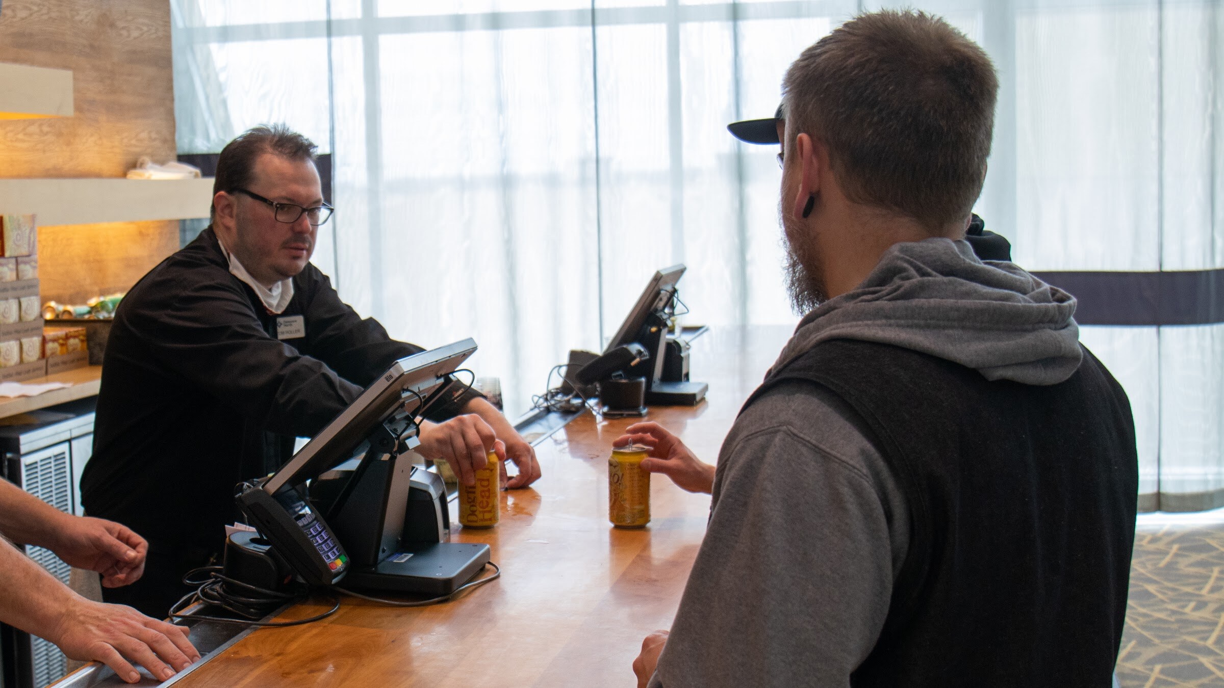 A bartender in a black jacket and glasses sets a yellow can on a wooden bar with a card reader, while a bearded guest in a gray hoodie and black vest waits in front of sheer curtains.