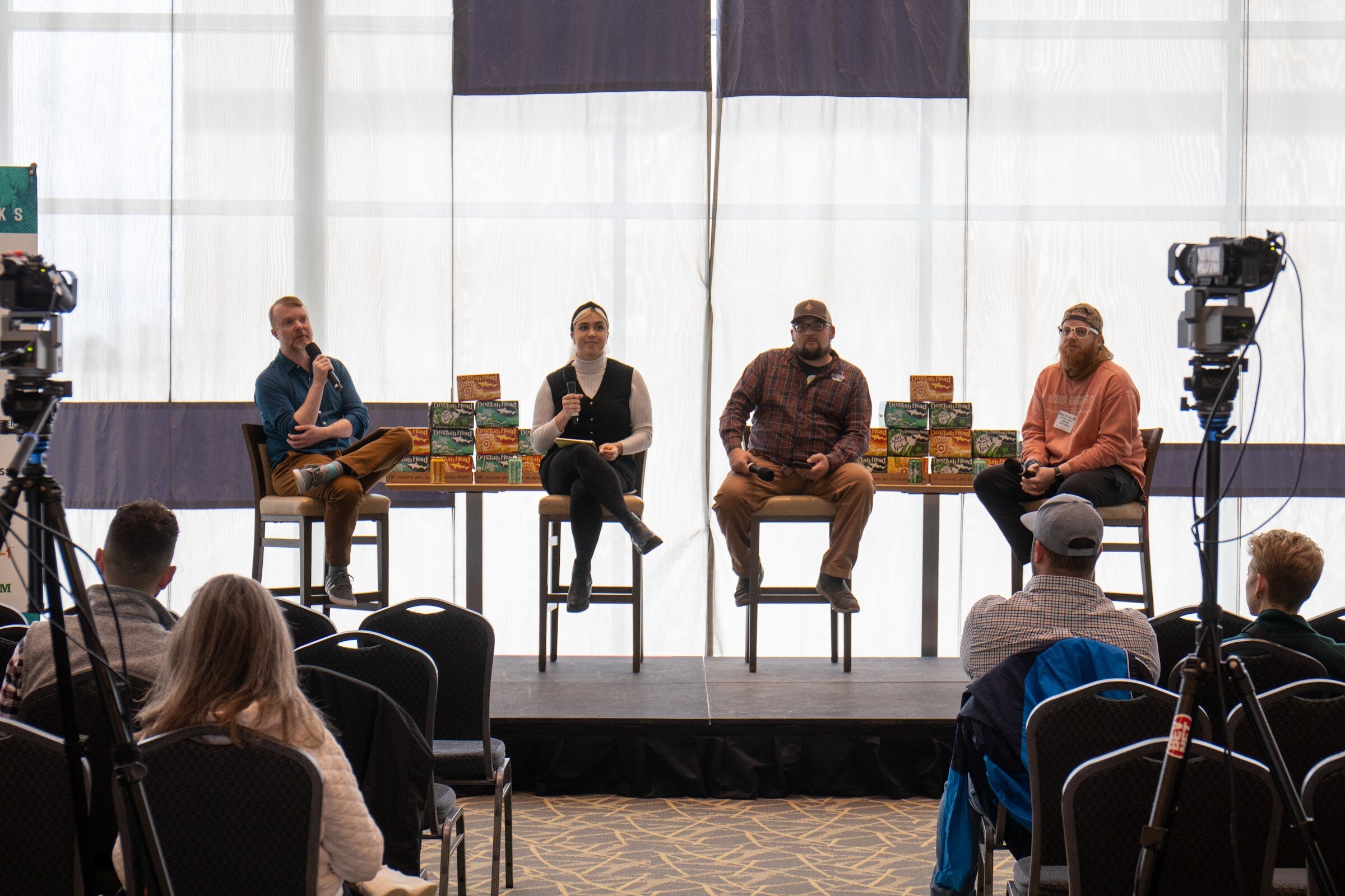Four people on tall stools on a low stage in front of sheer windows with Dogfish Head cartons stacked on the table between them, video cameras on tripods at each side and audience members in the foreground.