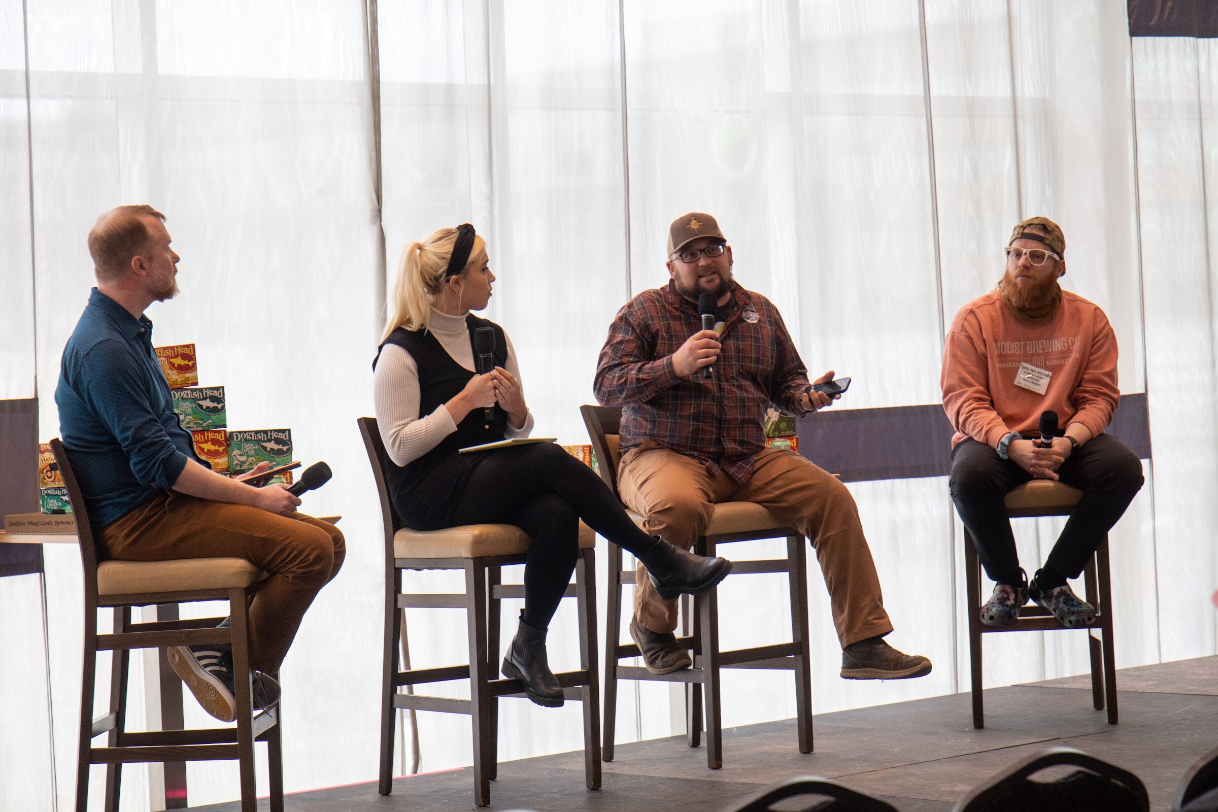 A man in a blue shirt and tan pants with a microphone, a woman with a black headband and vest, a bearded man in a plaid shirt and cap speaking into a microphone and a bearded man in an orange sweatshirt sit on stools beside stacks of Dogfish Head cartons.