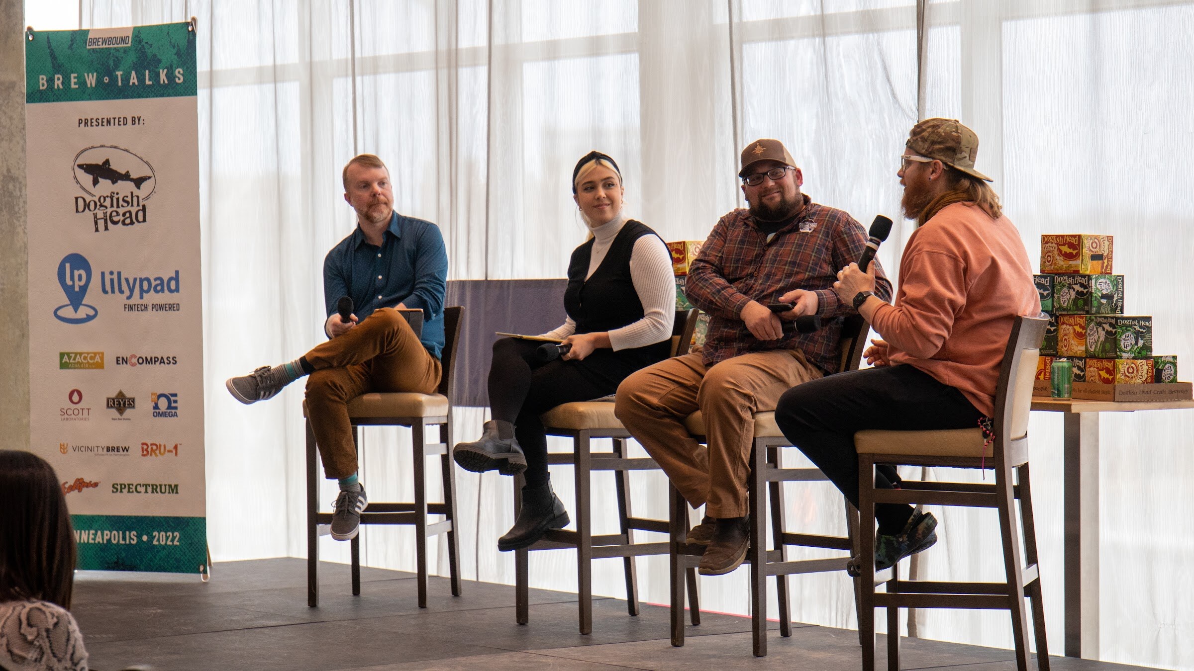 A bearded man in an orange sweatshirt and camo cap speaks into a microphone while a man in a blue shirt, a woman with a black headband and a man in a plaid shirt and cap listen, beside a banner reading Brew Talks, presented by Dogfish Head and Lilypad Fintech Powered, Minneapolis 2022.