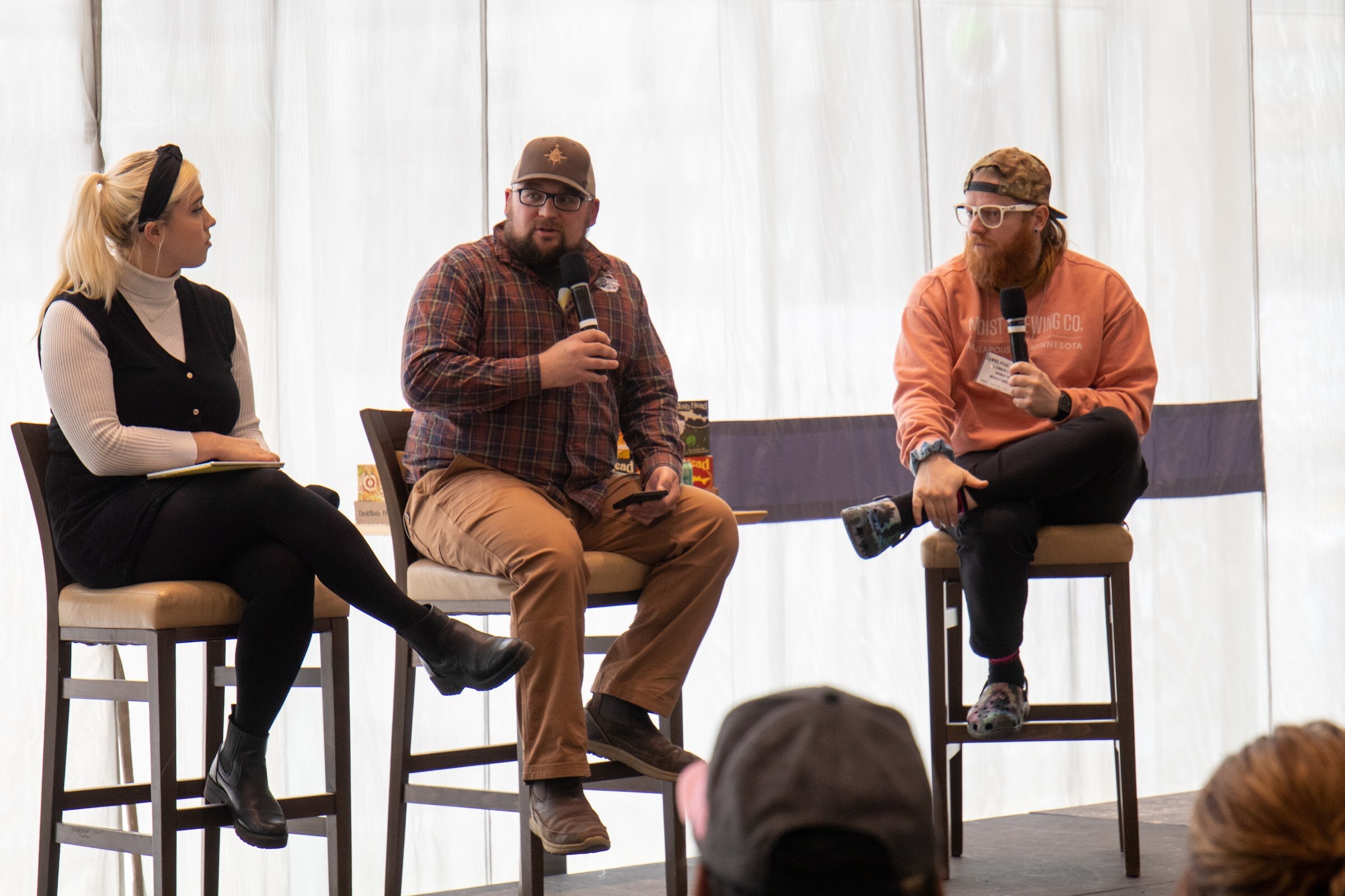 A woman in a black headband and vest holds a notebook and looks toward a bearded man in a plaid shirt and cap and a bearded man in an orange sweatshirt, both holding microphones, on stools in front of sheer curtains.
