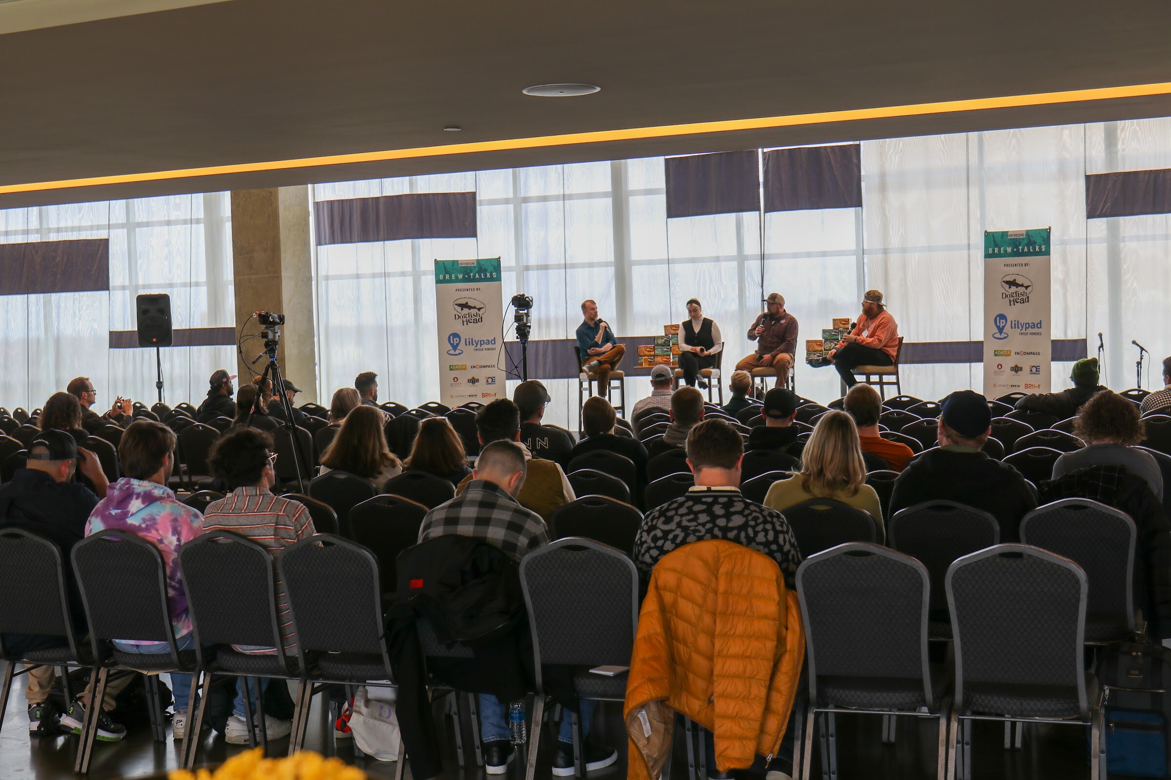 Rows of attendees seen from behind in black chairs, one with an orange puffer jacket on the seat, facing four panelists on a stage between two Brew Talks banners and a video camera on a tripod.