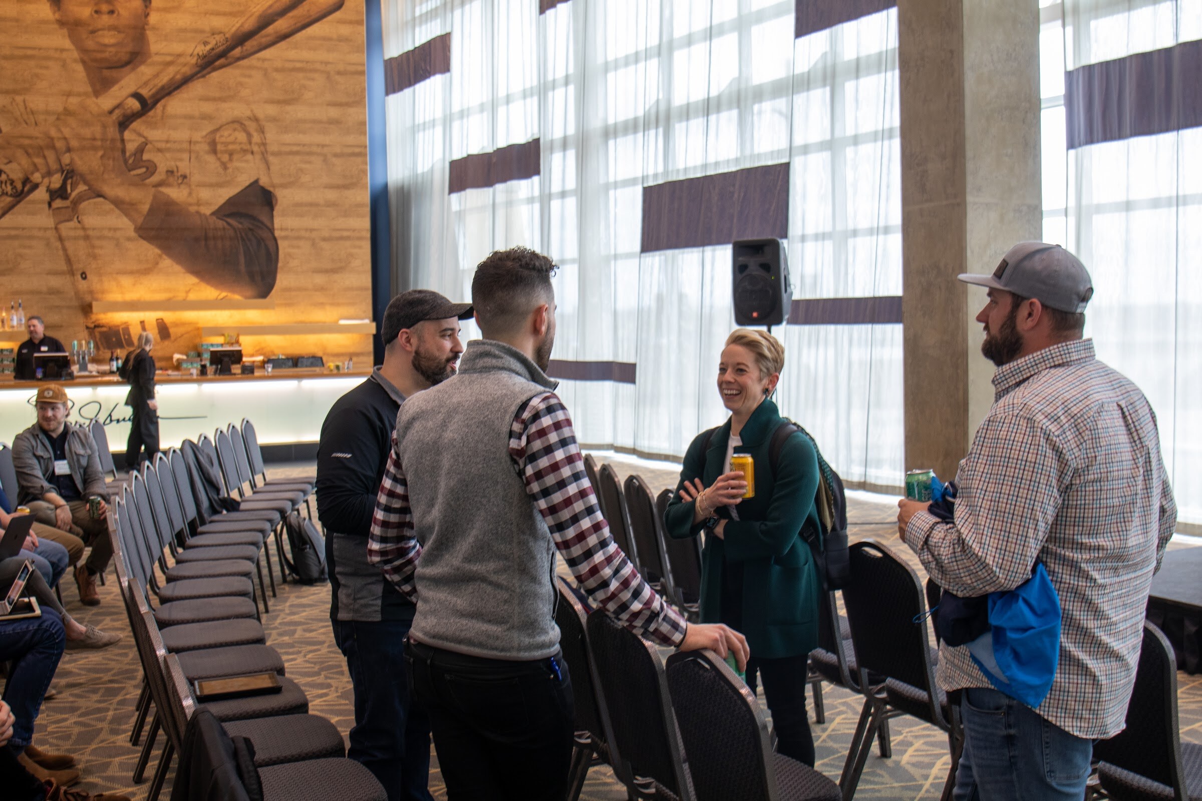 A man in a plaid shirt and gray vest, a man in a dark cap and black jacket, a smiling woman in a green jacket and a man in a gray cap and checked shirt talk beside rows of black chairs, with a large wood-toned mural of a batter on the wall.