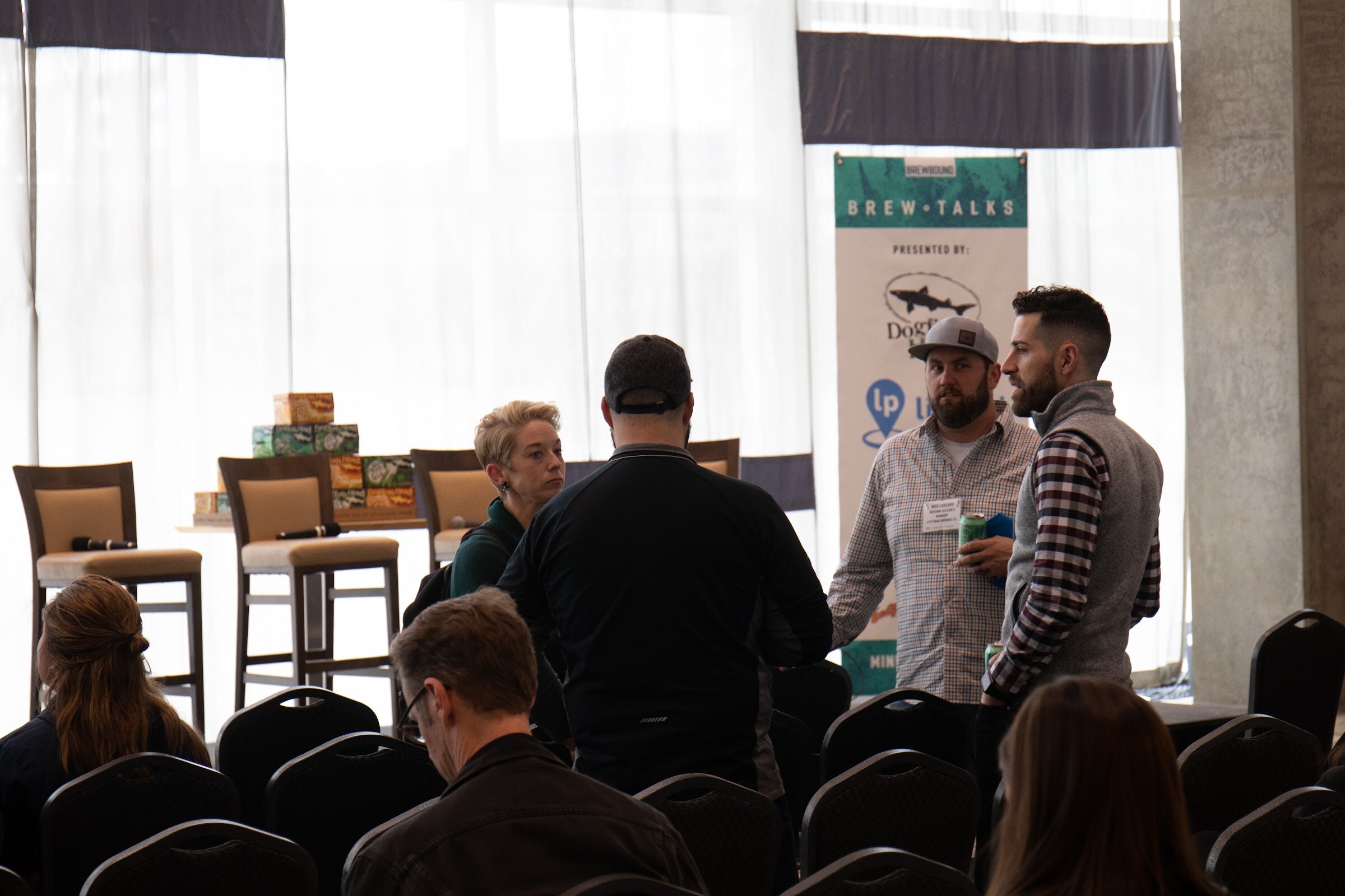 A small group of guests talk in front of tall bar stools and a table stacked with Dogfish Head cartons, with a Brew Talks banner behind them and rows of black chairs in the foreground.
