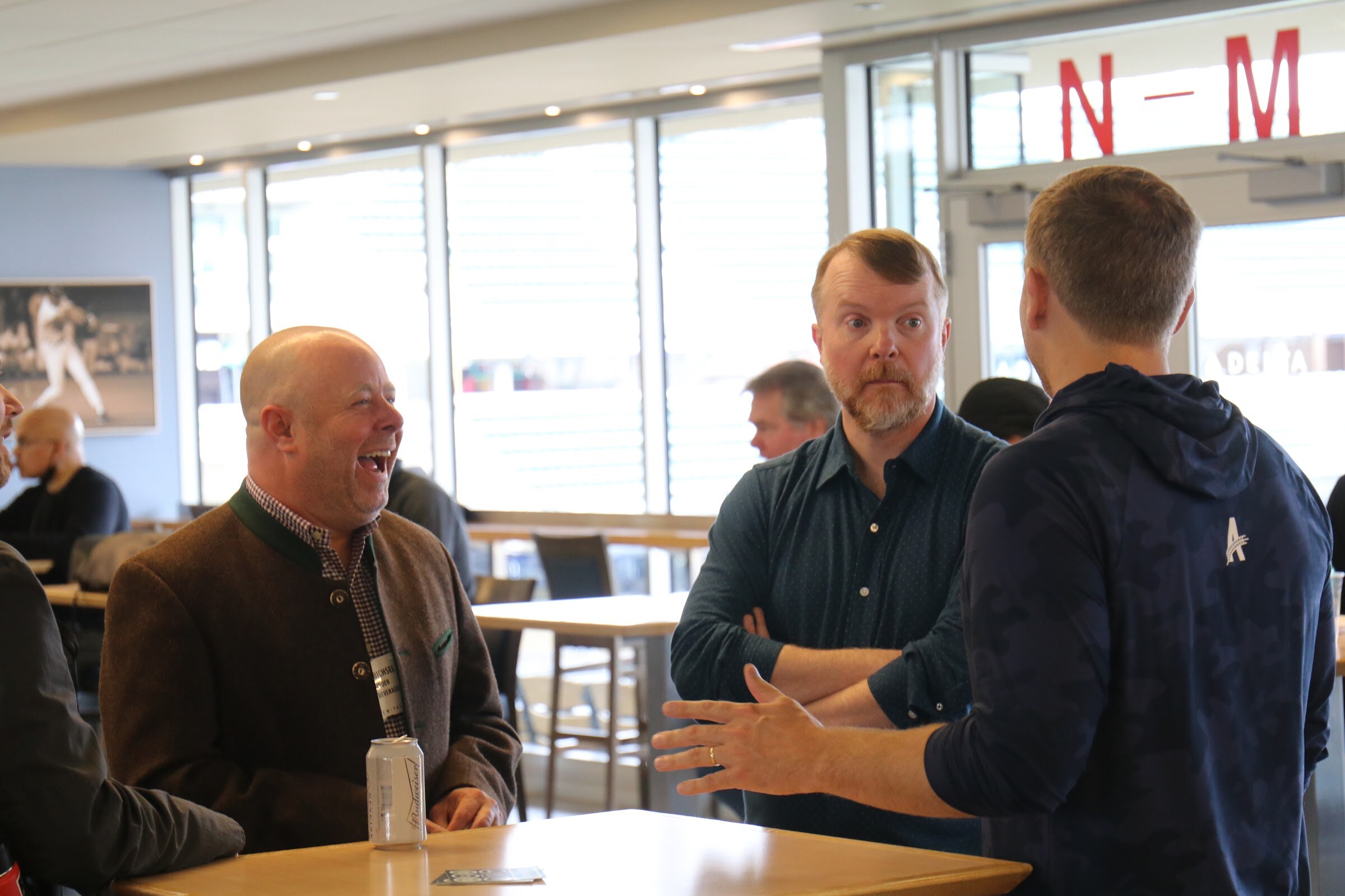 A laughing bald man in a brown jacket beside a can on a high-top table, a bearded man in a dark shirt with folded arms, and a man in a navy hoodie with a small logo gesturing, in front of tall windows.