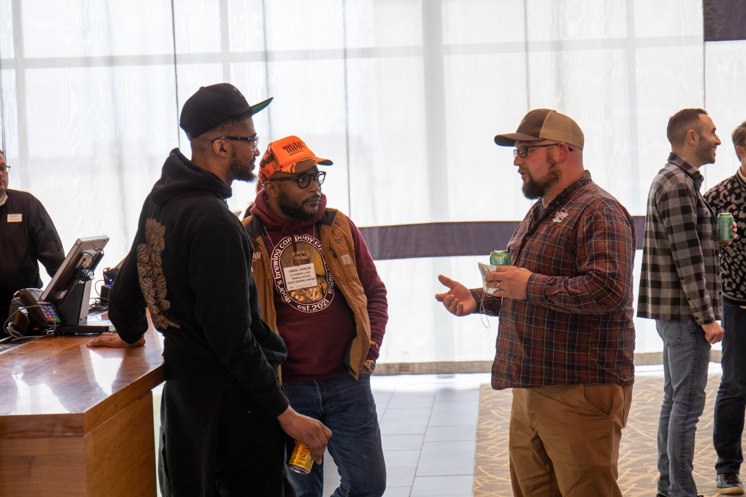A man in a black hoodie and cap, a man in an orange cap and maroon sweatshirt with a name badge, and a bearded man in a plaid shirt and tan cap talk, holding cans, beside a wooden bar counter in front of sheer curtains.