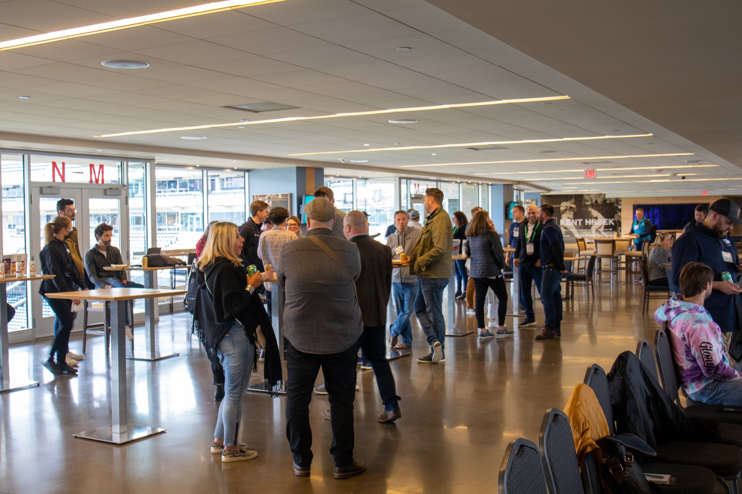 Wide view of guests standing and talking with canned drinks around tall tables in a long room with large windows, with rows of black chairs in the foreground right and a Kent Hrbek photo wall at the far end.