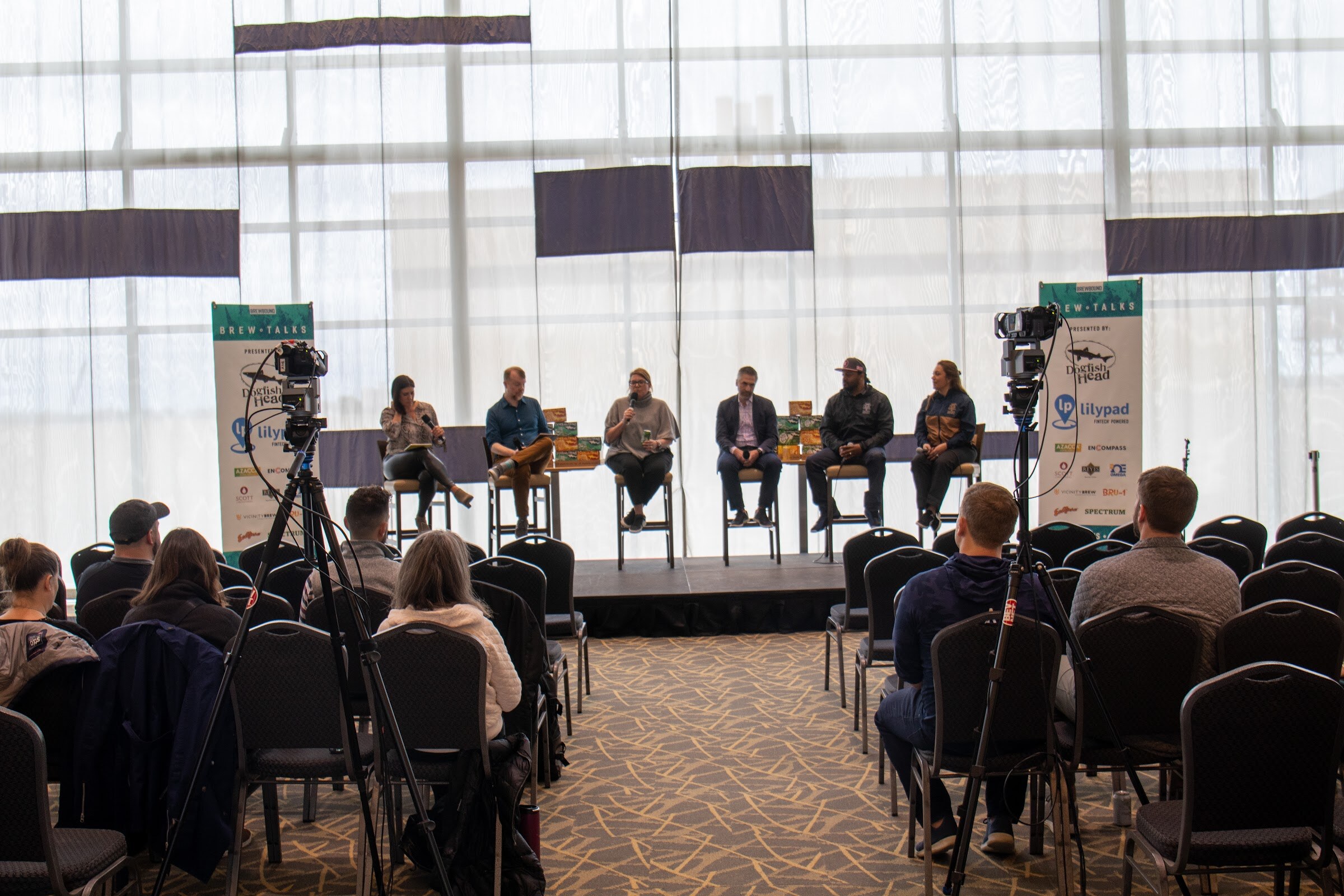 Six people on tall stools on a stage between two Brew Talks banners with Lilypad and Dogfish Head logos, with a video camera on a tripod at each side and seated attendees in black chairs in the foreground.