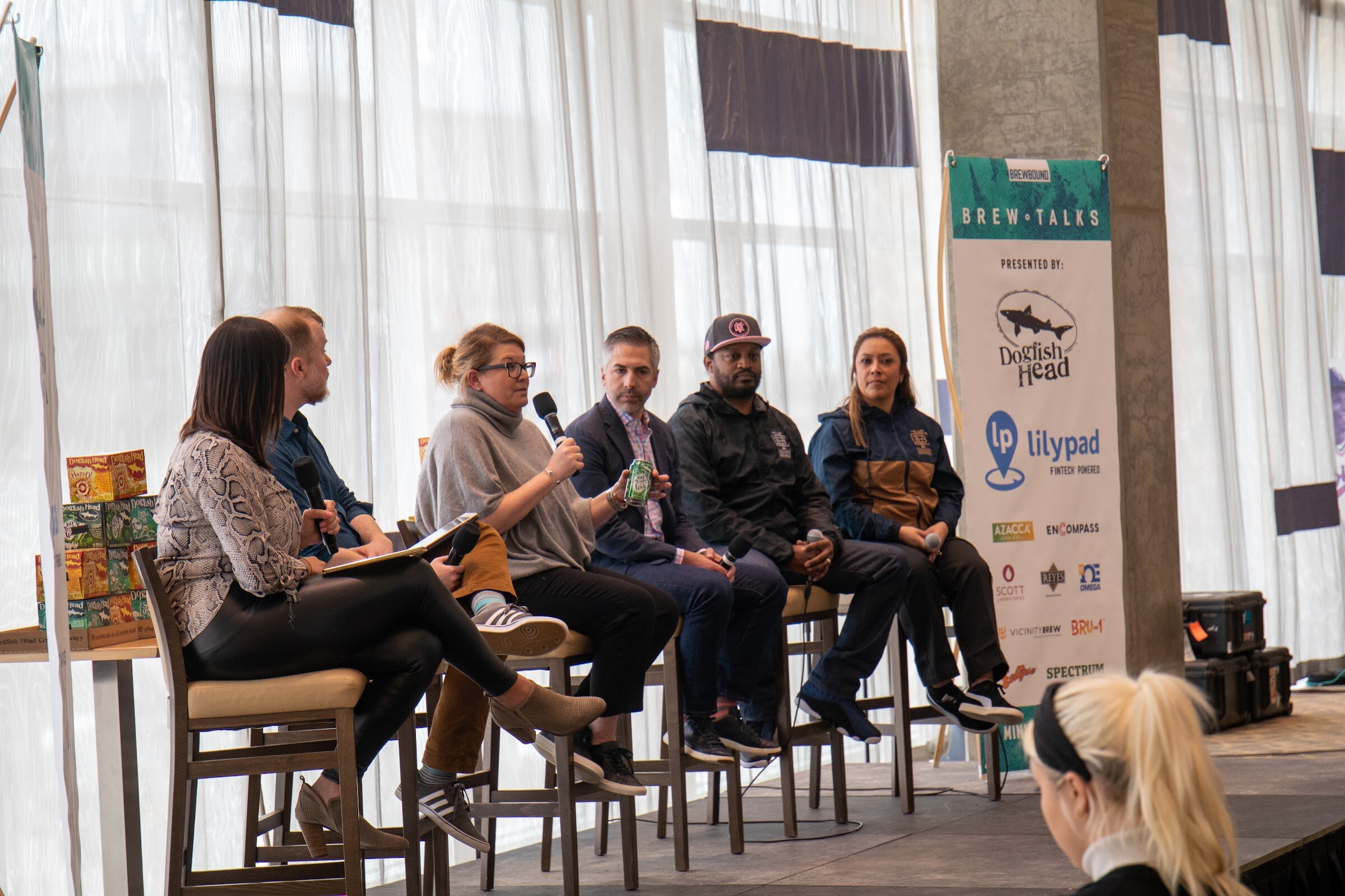 A woman in a gray cowl-neck sweater and glasses holds a green can and a microphone while a man in a navy blazer, a man in a black jacket and a woman in a navy and tan jacket sit beside her, with a woman in a snakeskin print top at left, next to a Brew Talks banner.