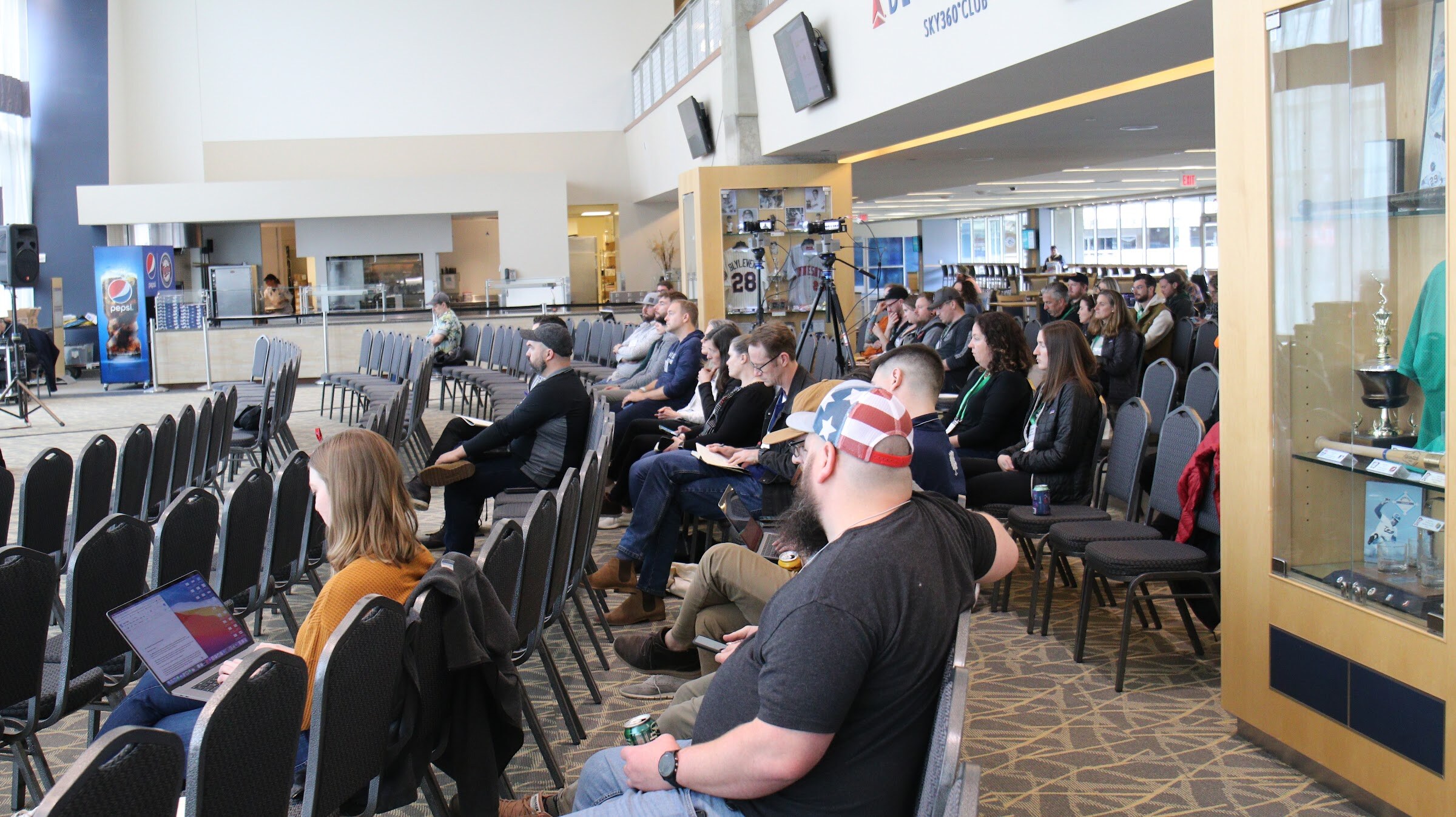 Seated attendees in rows of black chairs, including a bearded man in an American flag cap and a woman with a laptop, in a large hall with a Pepsi cooler, a food counter and a trophy case at the right.