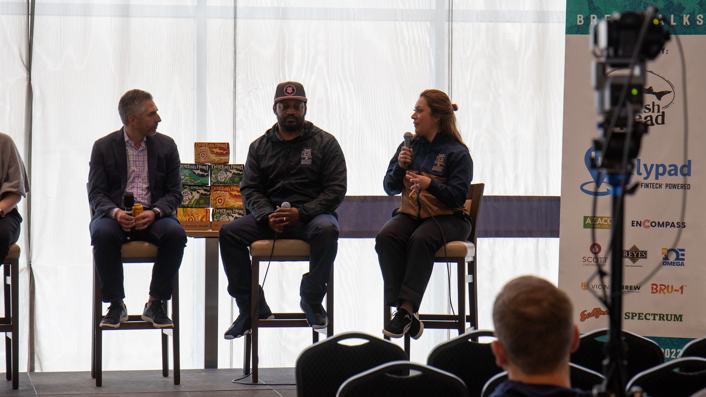 A man in a navy blazer and pink checked shirt, a man in a dark cap and black jacket and a woman in a navy and tan jacket speaking into a microphone sit on tall stools beside a stack of Dogfish Head cartons and a Lilypad banner at the right.