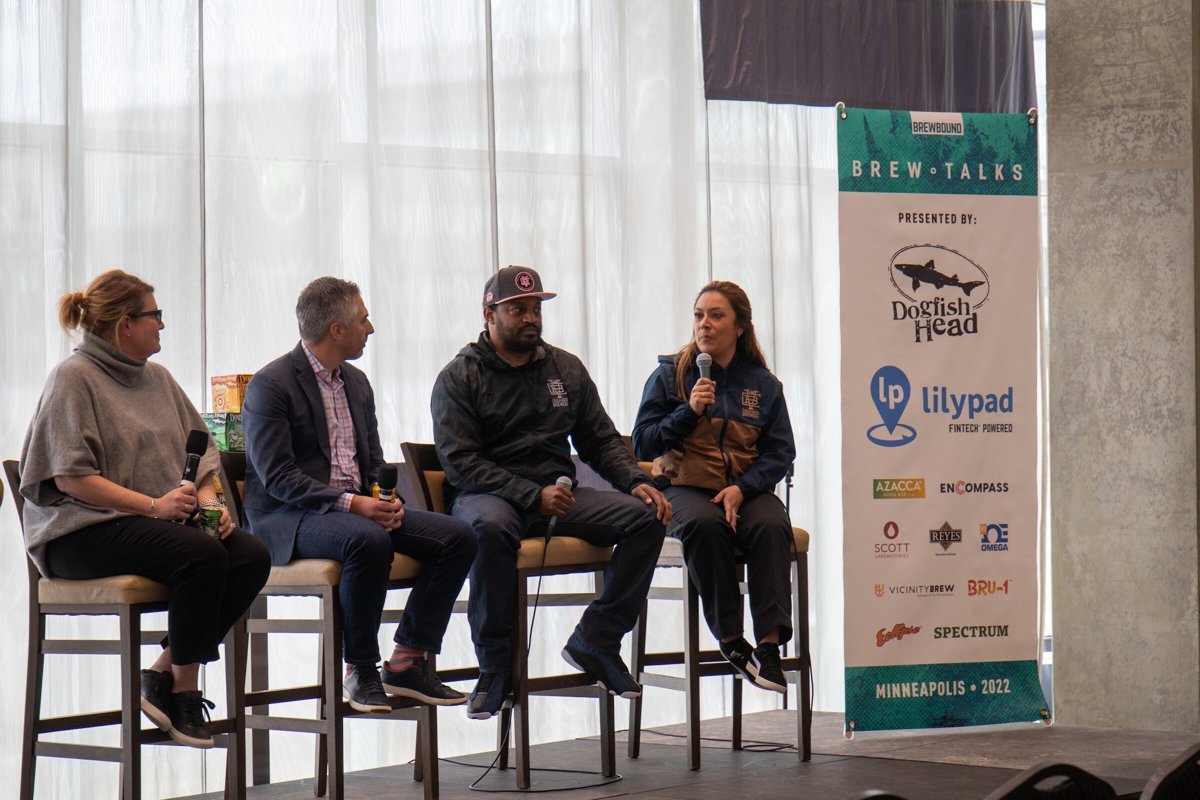 A woman in a navy and tan jacket speaks into a microphone beside a man in a dark cap and black jacket, a man in a navy blazer and a woman in a gray sweater on stools, with a Brew Talks banner listing Dogfish Head and Lilypad Fintech Powered behind them.