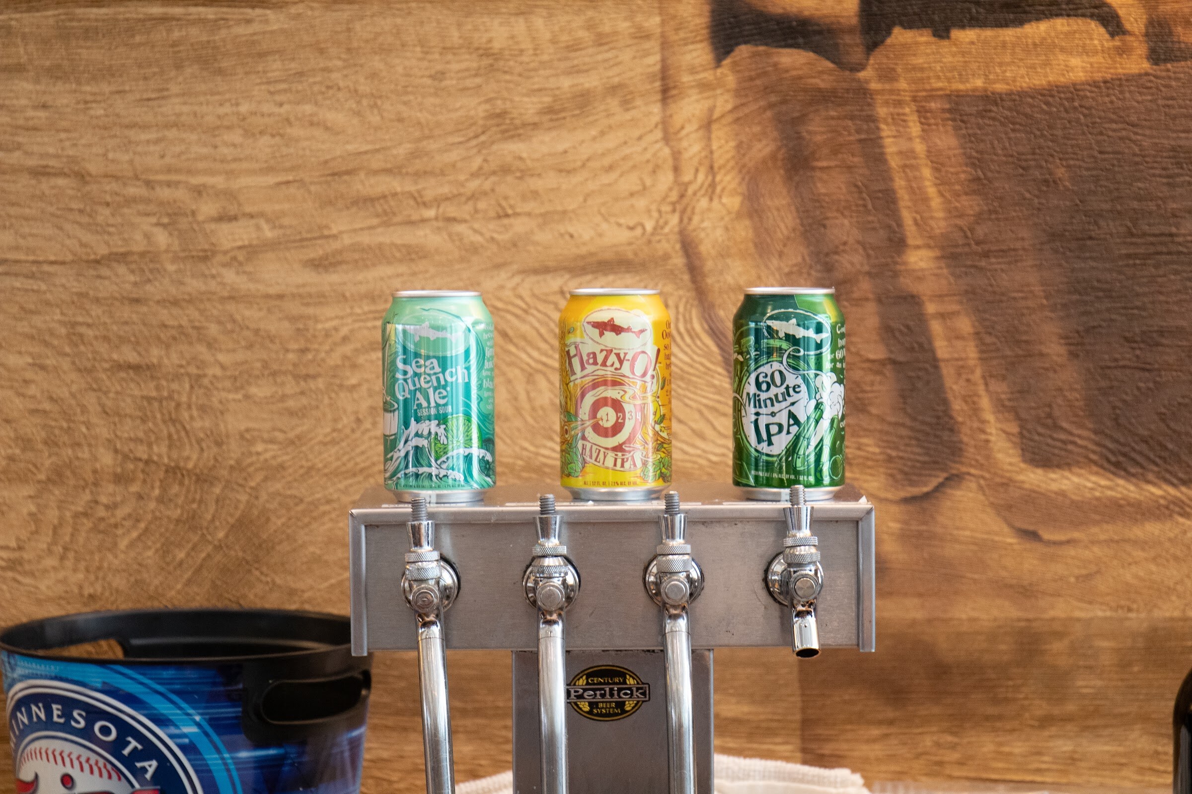 Three Dogfish Head cans, Sea Quench Ale, Hazy-O! and 60 Minute IPA, sit on a chrome four-tap tower in front of a wood-grain wall, beside a blue Minnesota Twins bucket.