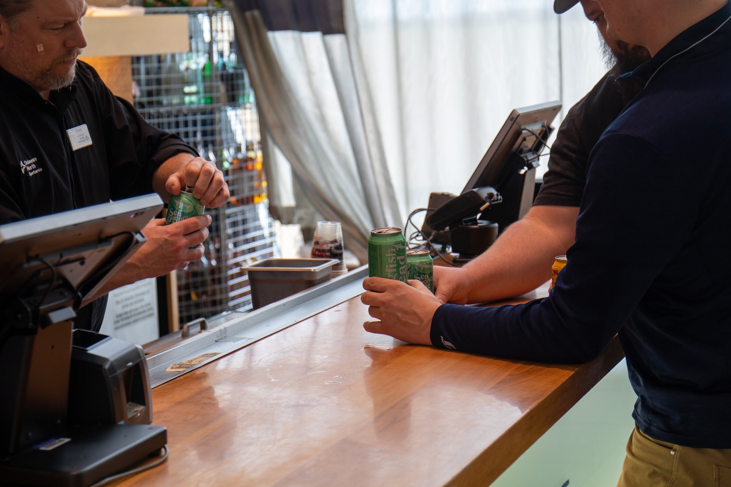 A bartender in a black polo opens a green can behind a register while a guest in a navy long-sleeve shirt rests his hands on two green cans on a wooden bar, in front of sheer curtains.