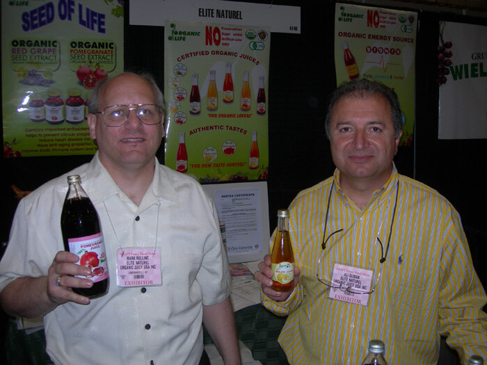 Mark Rollino of Elite Naturel, left, holds a bottle of pomegranate juice next to another man holding a juice bottle, in front of Elite Naturel and Seed of Life posters.