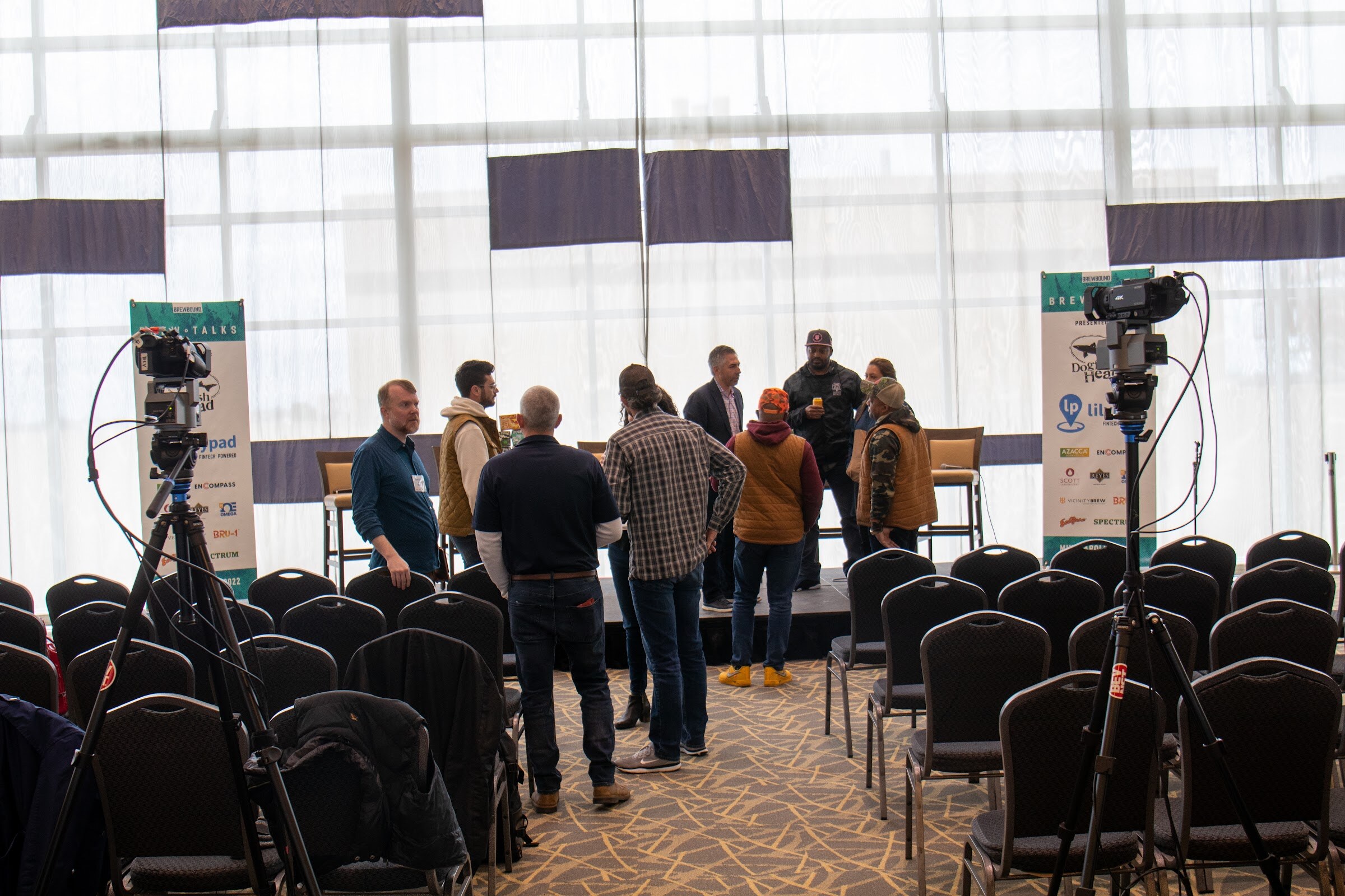 A group of about eight people stand and talk in front of a small stage with tall bar stools, between two video cameras on tripods and two Brew Talks banners with Lilypad and Dogfish Head logos, with rows of black chairs in the foreground.