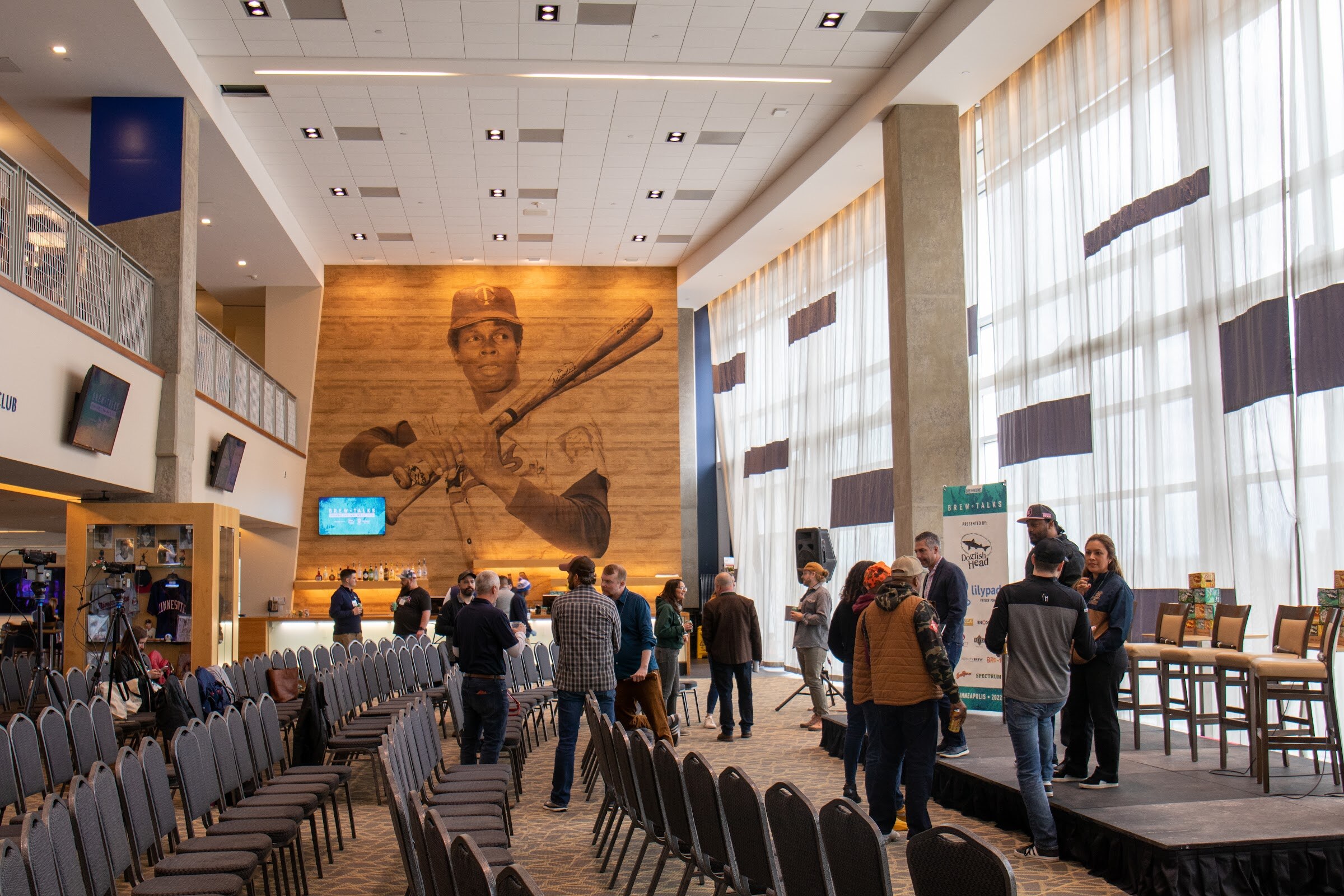 A tall hall with rows of black chairs, a small stage with bar stools and a Brew Talks banner at the right, a large wood-toned mural of a batter on the wall and guests standing and talking.