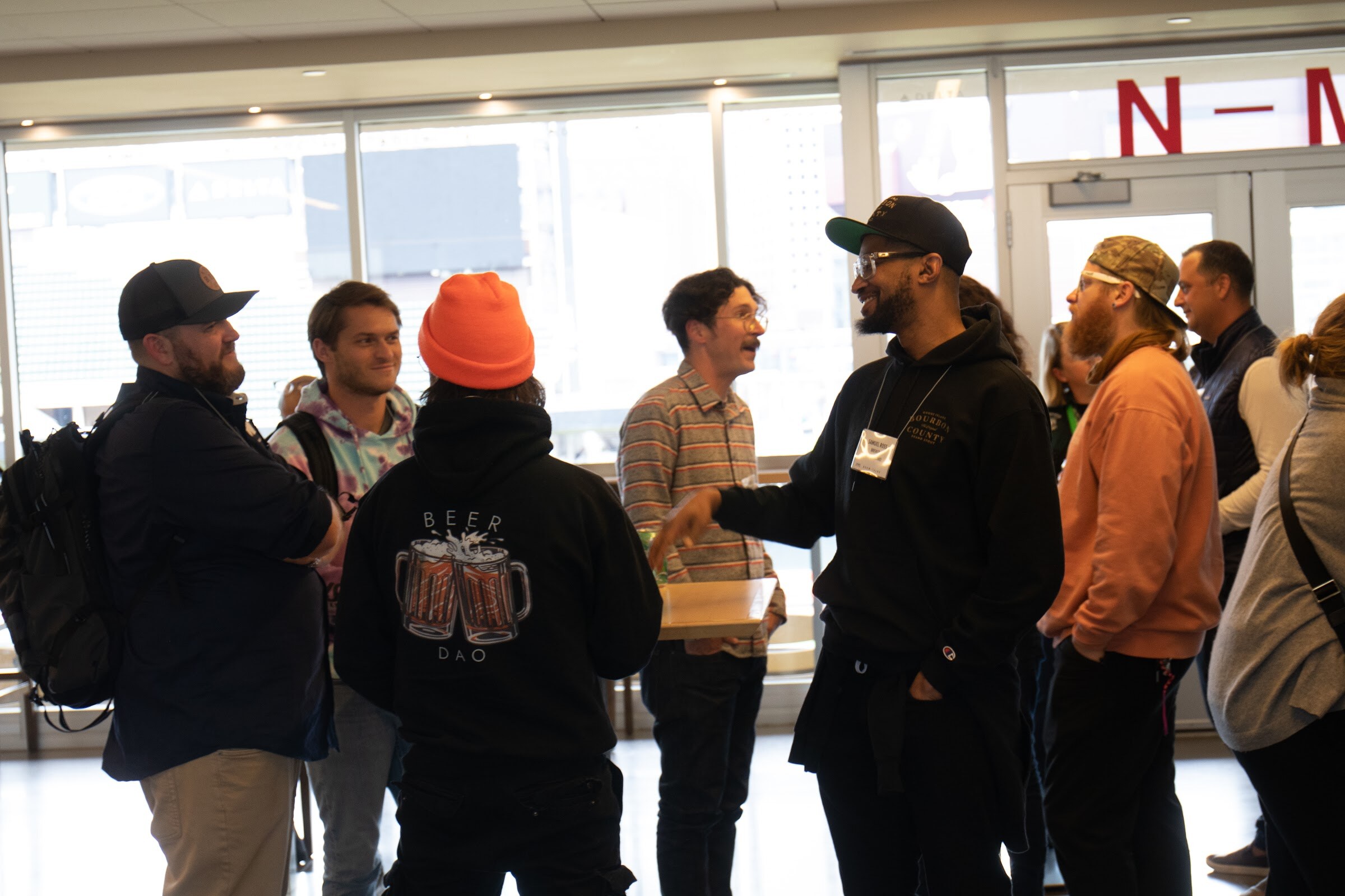 A group of guests talk and smile in front of tall windows, including a man in a black cap, a person in an orange beanie and a black Beer Dao hoodie seen from behind, and a man in glasses and a black hoodie gesturing.