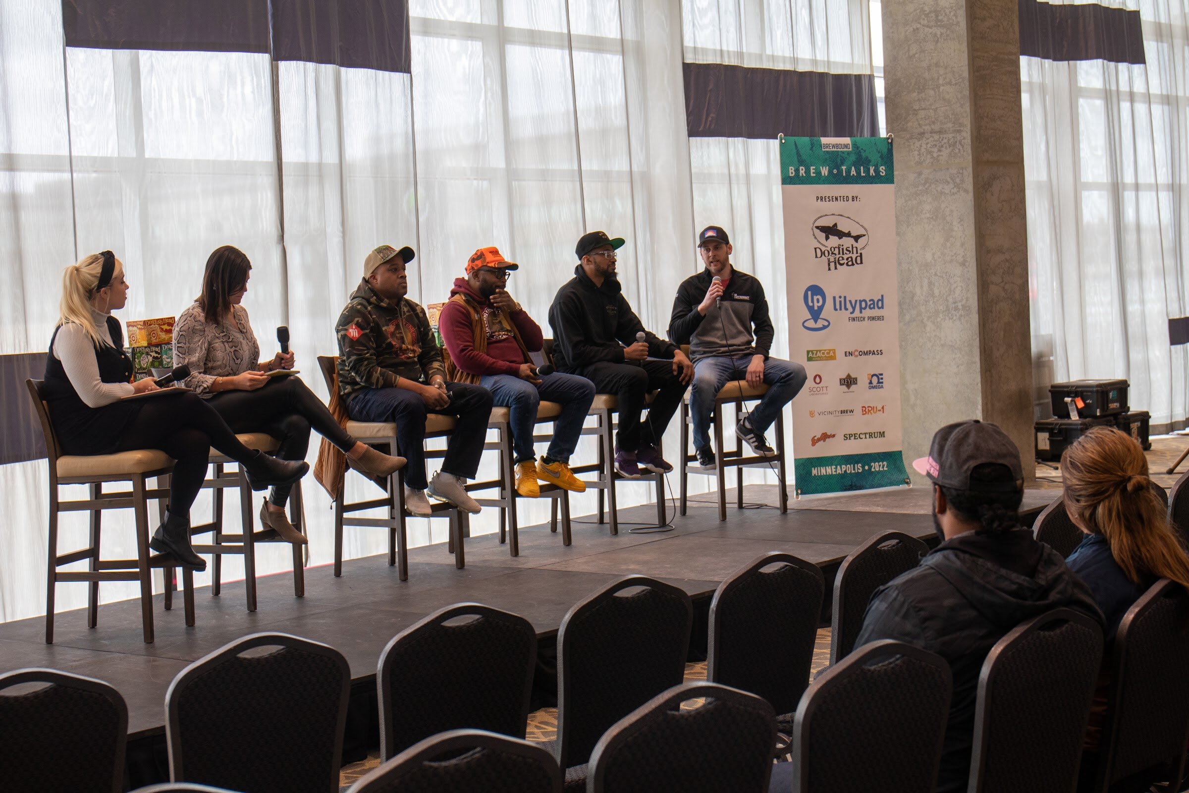 A woman with a black headband and a woman in a snakeskin print top hold microphones beside four men on tall stools, in a camo hoodie, an orange cap and maroon sweatshirt, a black hoodie and a gray quarter-zip speaking, next to a Brew Talks banner, with rows of black chairs in the foreground.