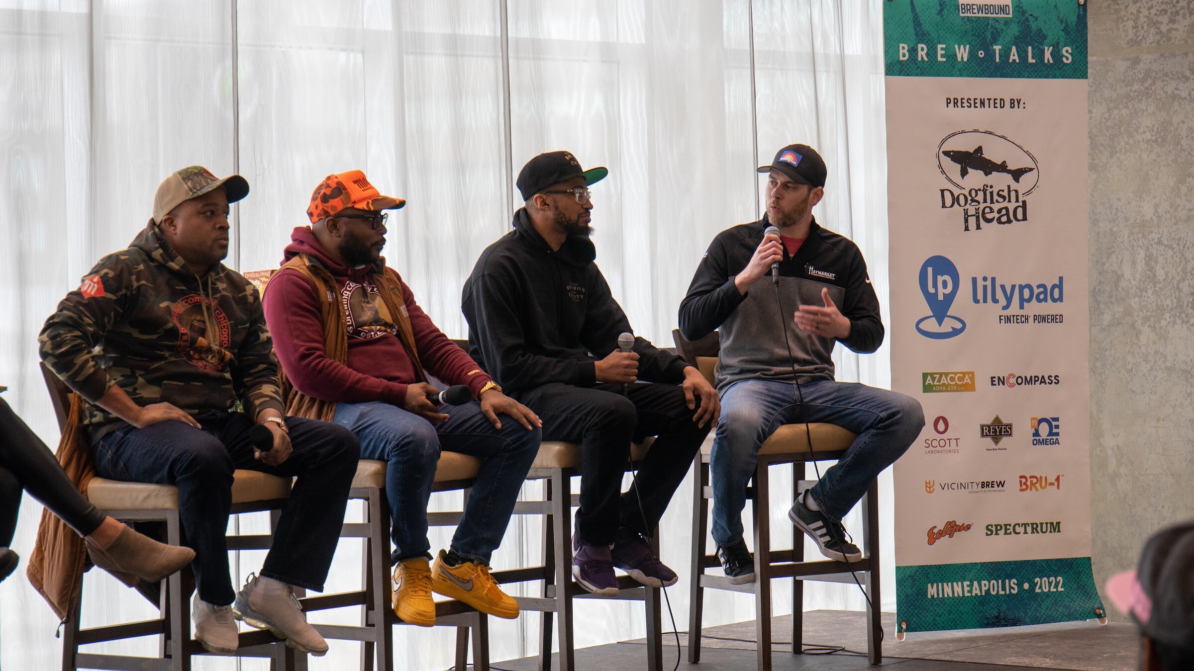 Four men in caps sit on stools in front of sheer curtains: a man in a camo hoodie, a man in an orange cap and maroon sweatshirt, a man in a black hoodie and a man in a gray quarter-zip speaking into a microphone, beside a Brew Talks banner.
