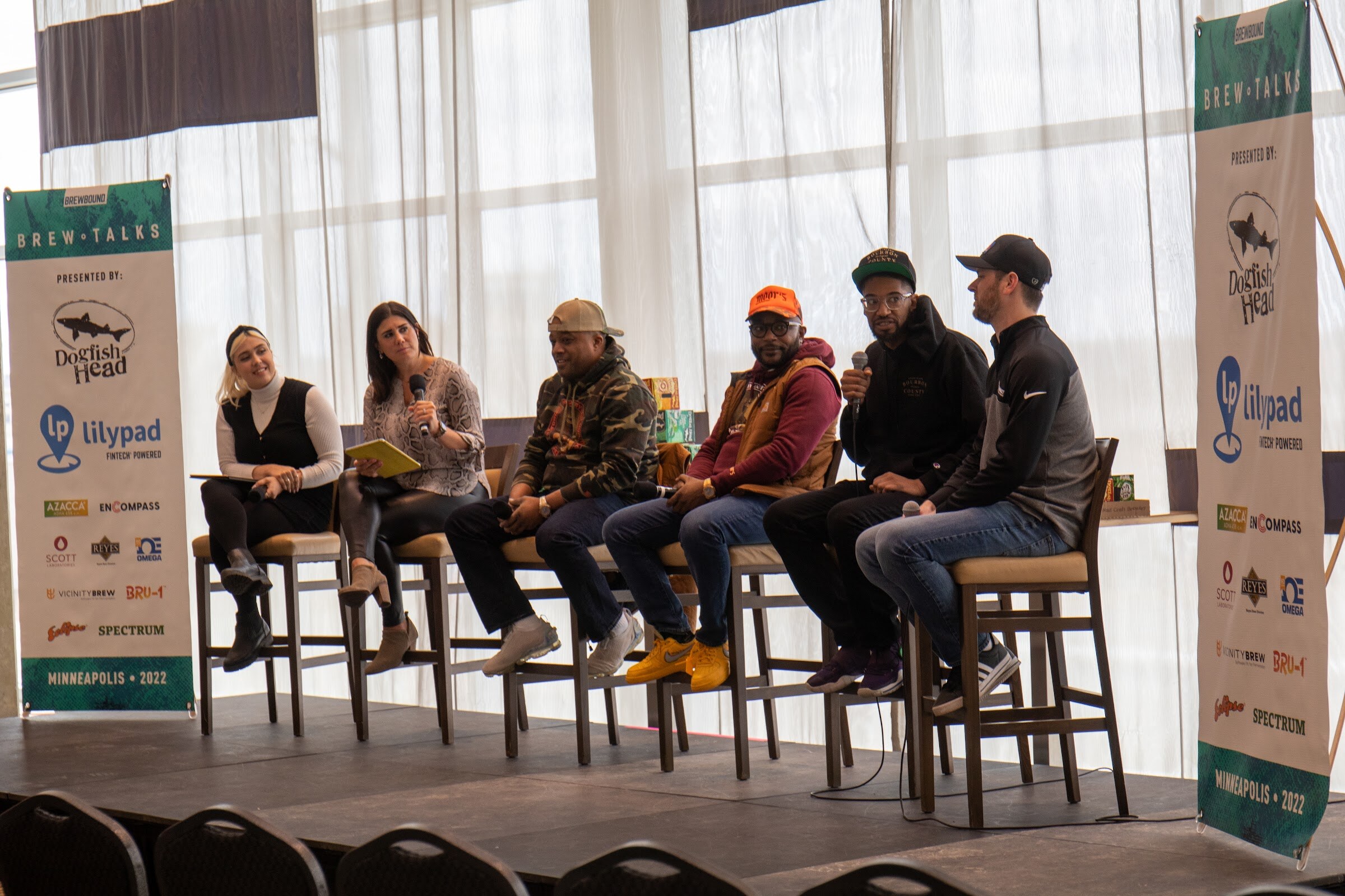 Six people on tall stools between two Brew Talks banners presented by Dogfish Head and Lilypad Fintech Powered, including a woman with a headband, a woman holding a yellow notebook and four men in caps, with black chairs in the foreground.