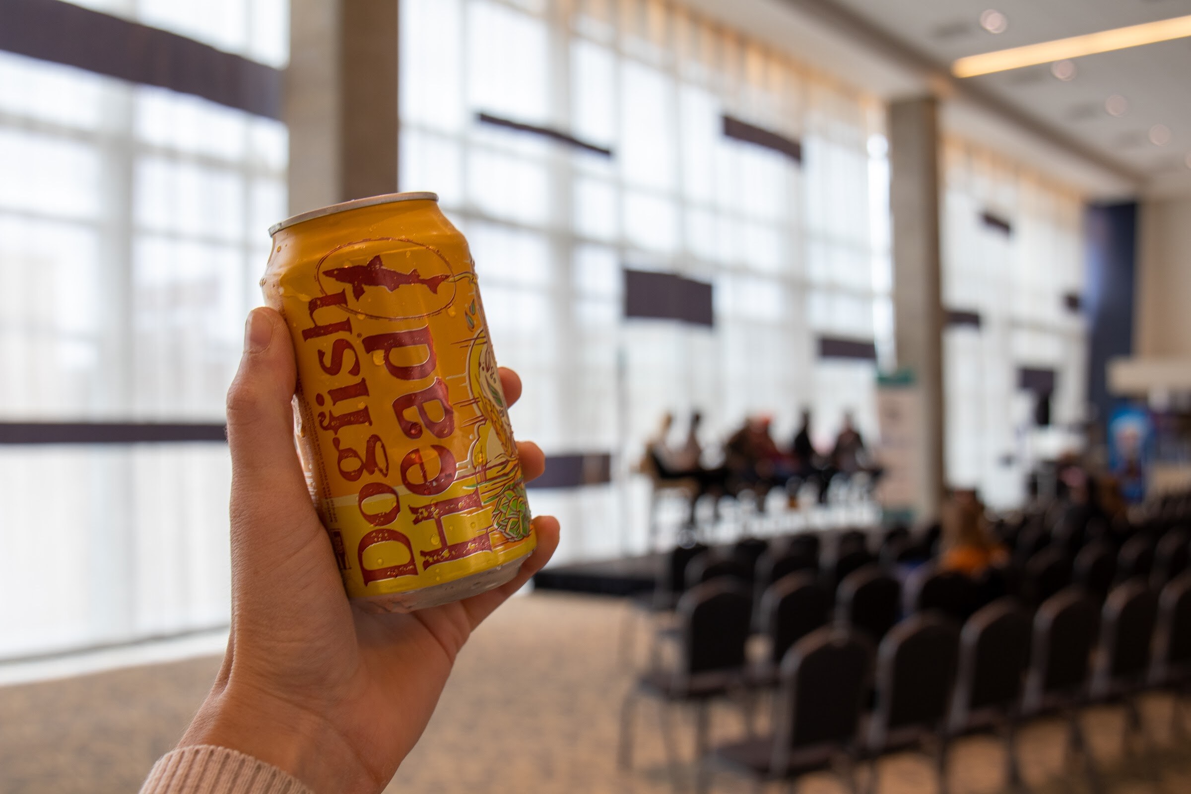 A hand holds a yellow Dogfish Head can in the foreground, with a blurred panel on a stage and rows of black chairs behind it in front of tall windows.
