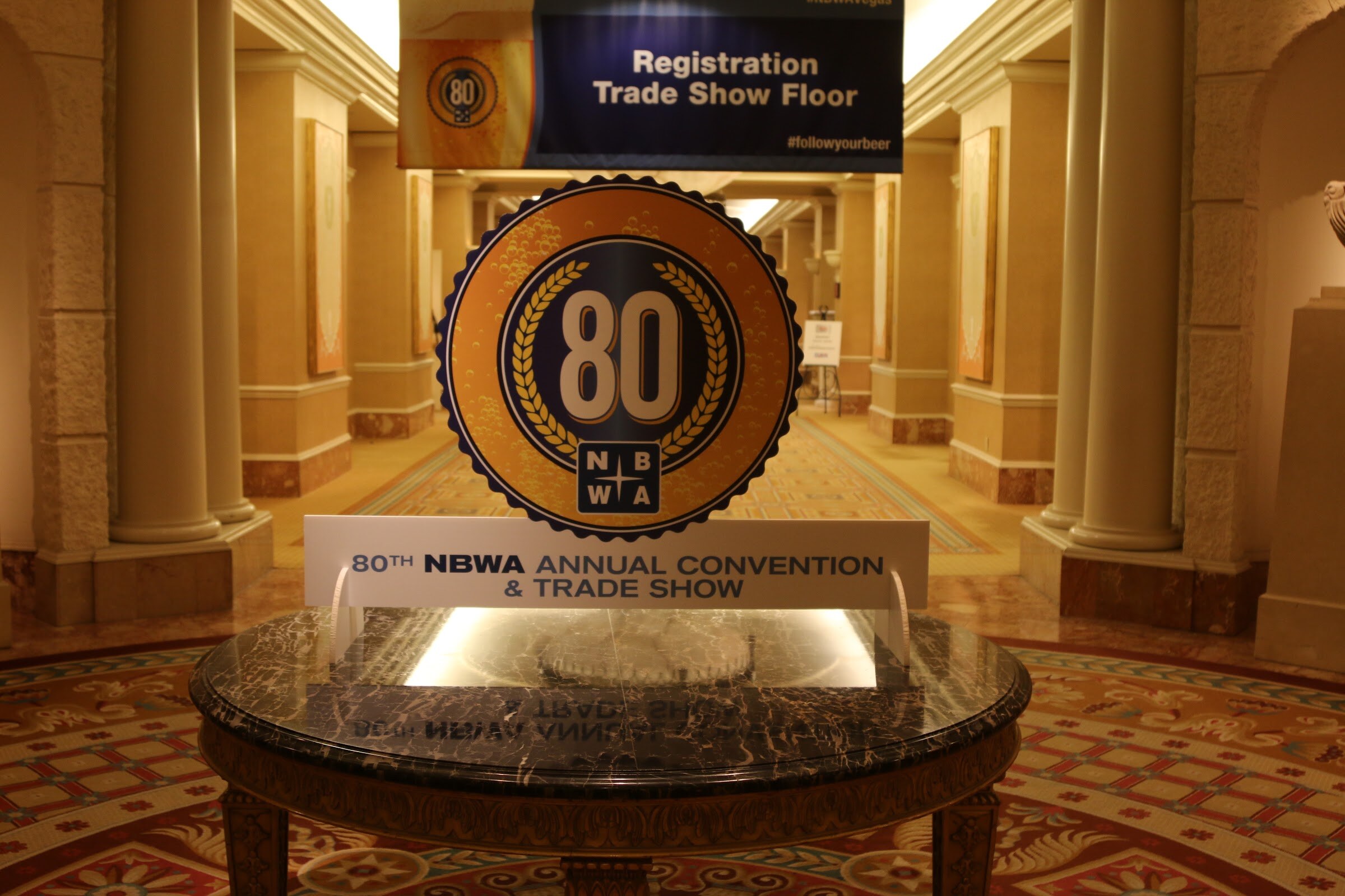 A round gold 80th anniversary NBWA emblem on a marble table beneath a Registration Trade Show Floor banner in an ornate hallway.
