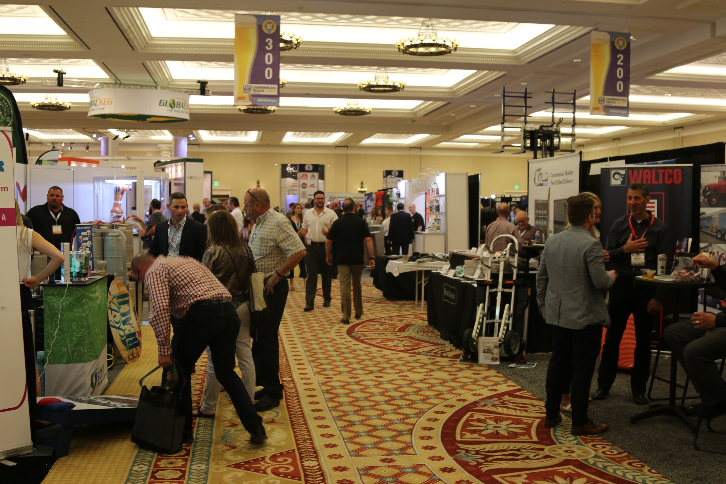 A carpeted trade show aisle with attendees walking and talking between booths, with Waltco signage and 200 and 300 aisle banners overhead.